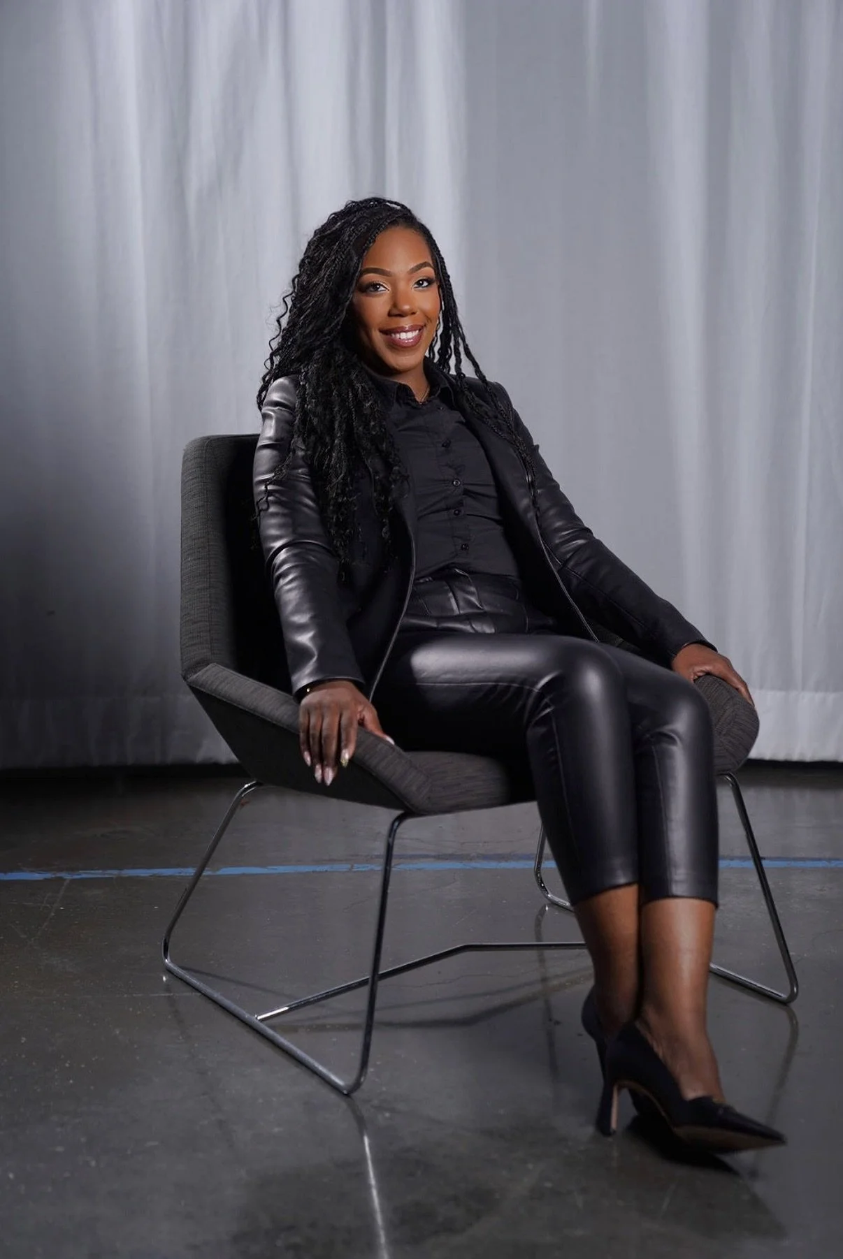 A confident woman with long braided hair sitting on a modern gray chair, smiling, wearing a black leather jacket, black shirt, black leather pants, and black heels in a minimal studio setting with a white curtain background.