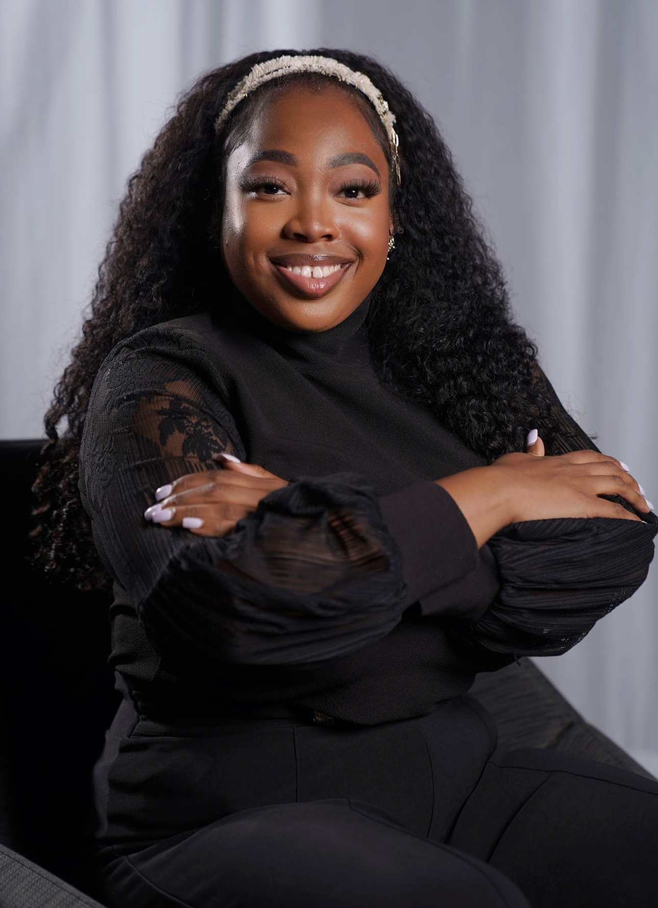 A smiling woman with curly hair, wearing a black top with lace sleeves and a headband, sitting with her arms crossed.