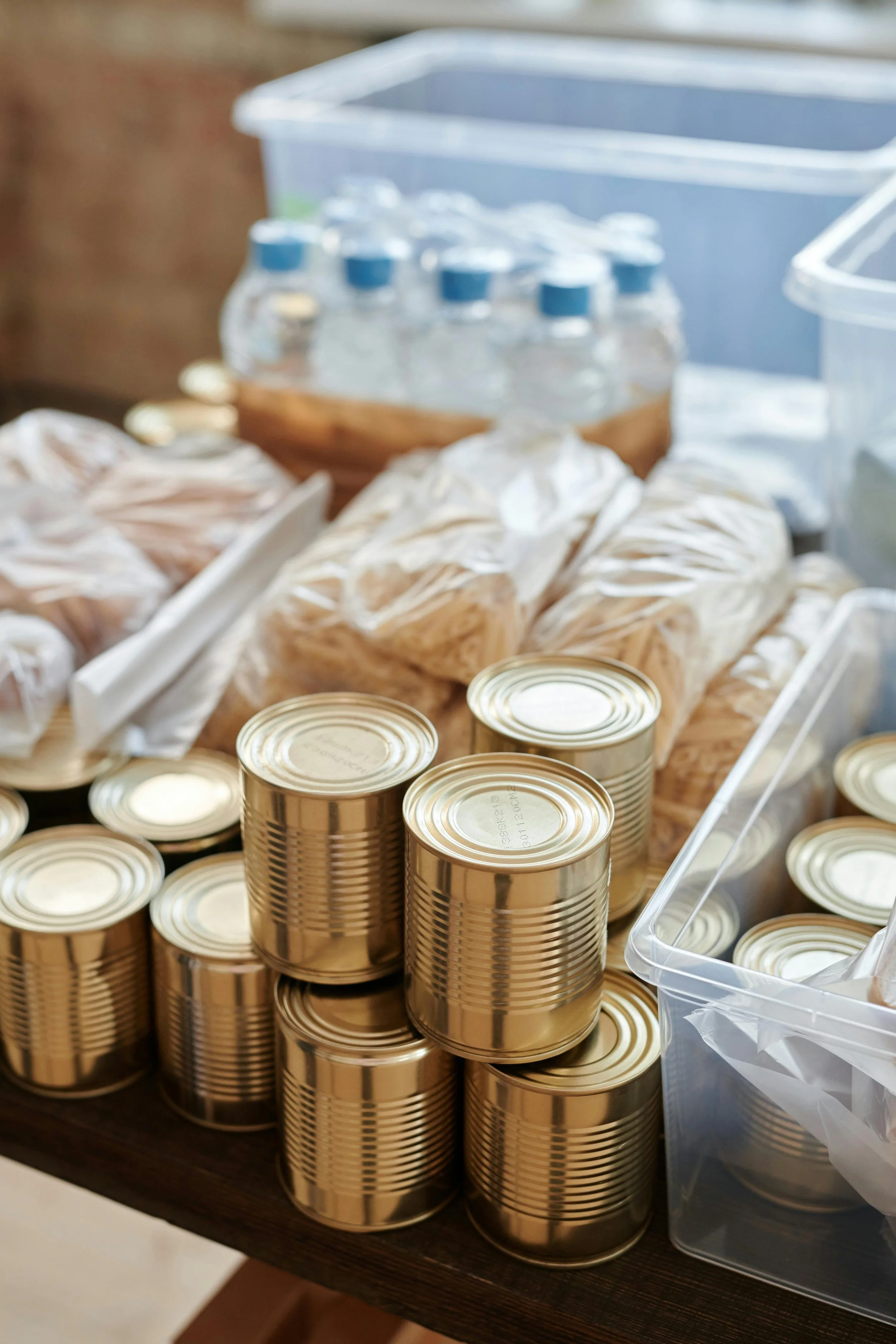 Multiple gold cans stacked on a brown wooden table with some bags of food and a clear plastic container nearby.