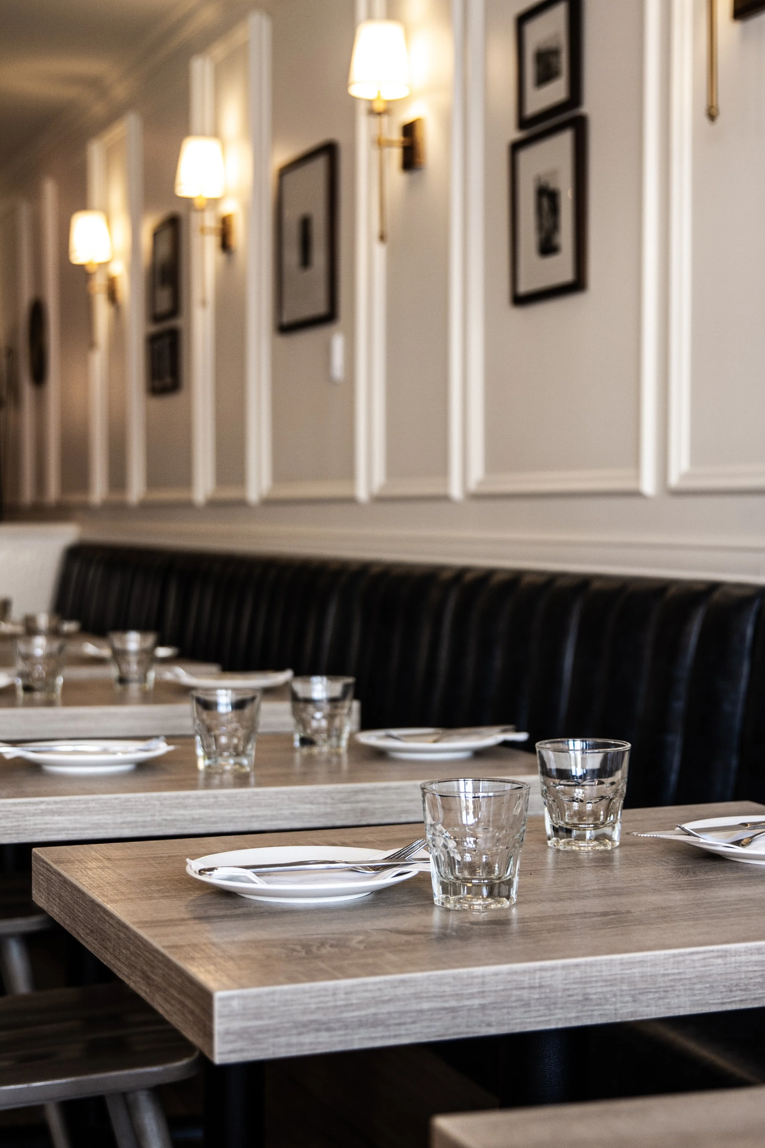 A dining area with wooden tables set with white plates, forks, and clear drinking glasses in front of black upholstered chairs. The background features a white wall with wainscoting decorated with framed black-and-white photos and warm wall sconces.