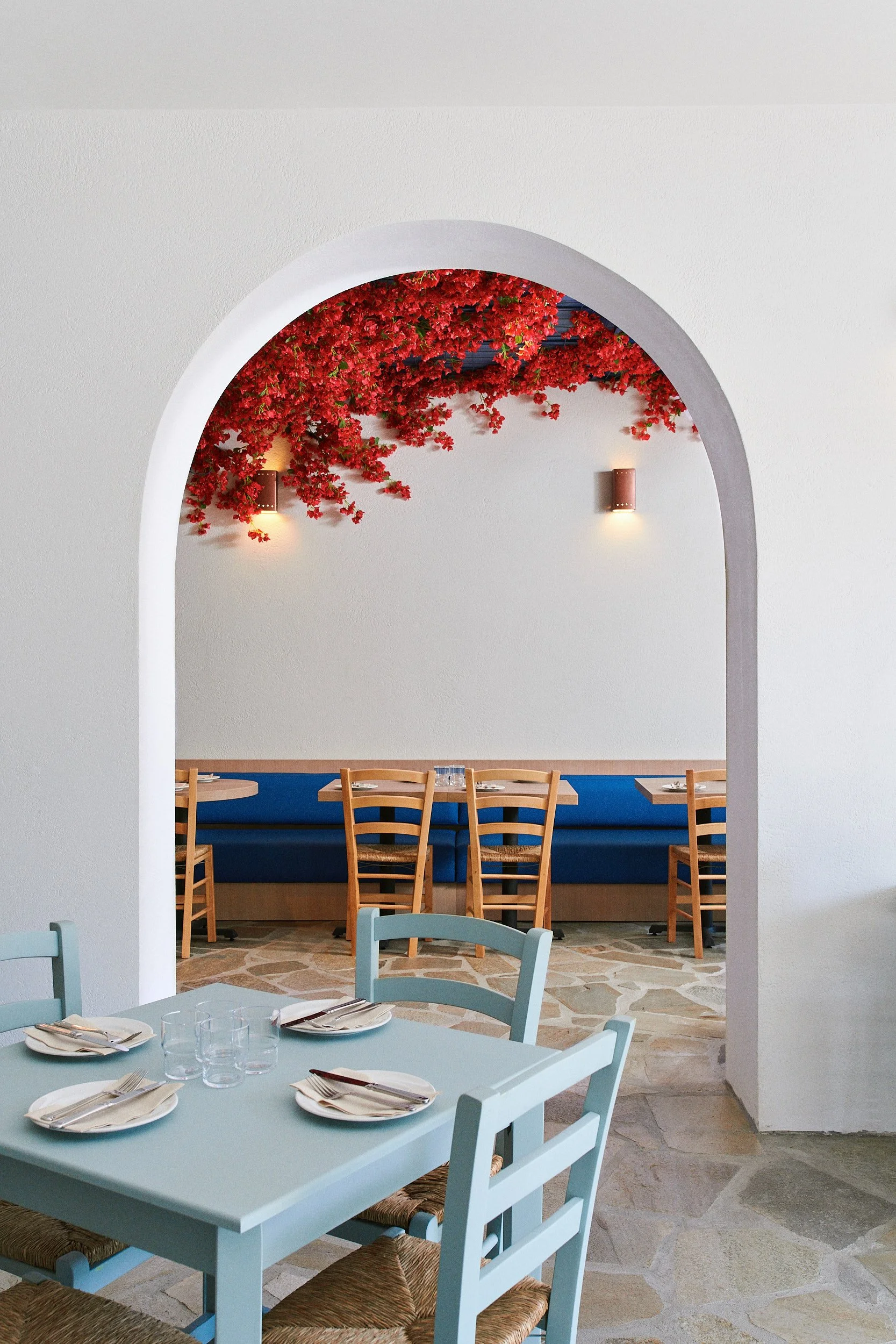 Dining area with a light blue table set with utensils and glasses, surrounded by matching chairs, with a stone floor, and an archway leading to a space with a ceiling decorated with red flowering vines and copper wall sconces on the back wall.
