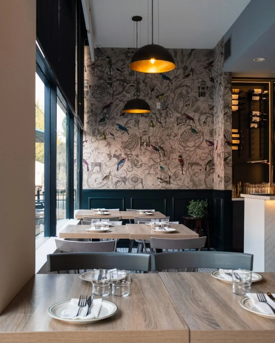 Sundrenched restaurant dining area with fully set wooden dining tables. A large window on the left side highlights decorative wallpaper with colorful bird illustrations. Brass pendant lights, and a bar with wine rack on the right.