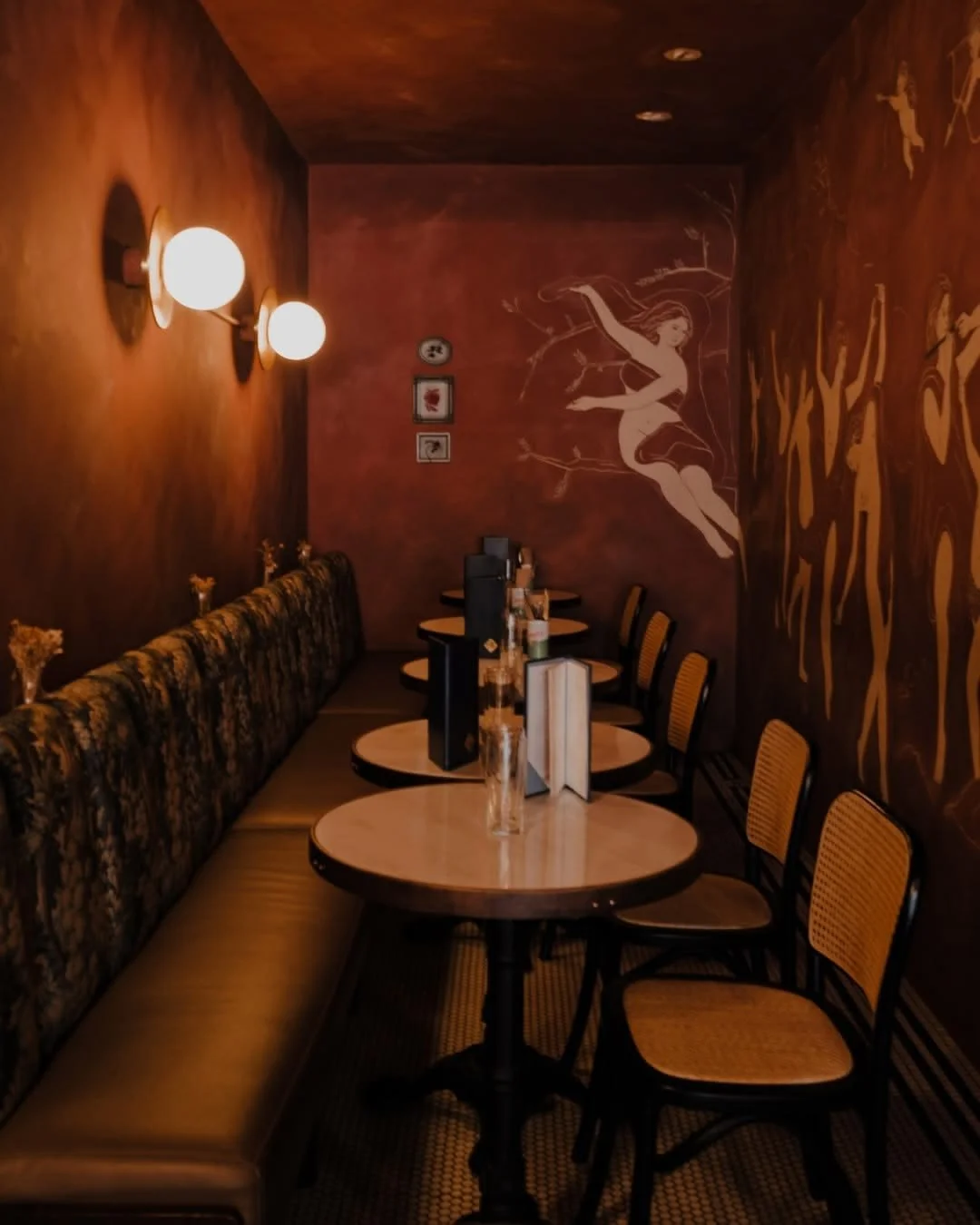 A cozy restaurant banquette with a long floral upholstered bench and four round tables set with glasses, menus, and condiments. The wall features a whimsical mural of women frolicking, with warm lighting from two wall sconces.