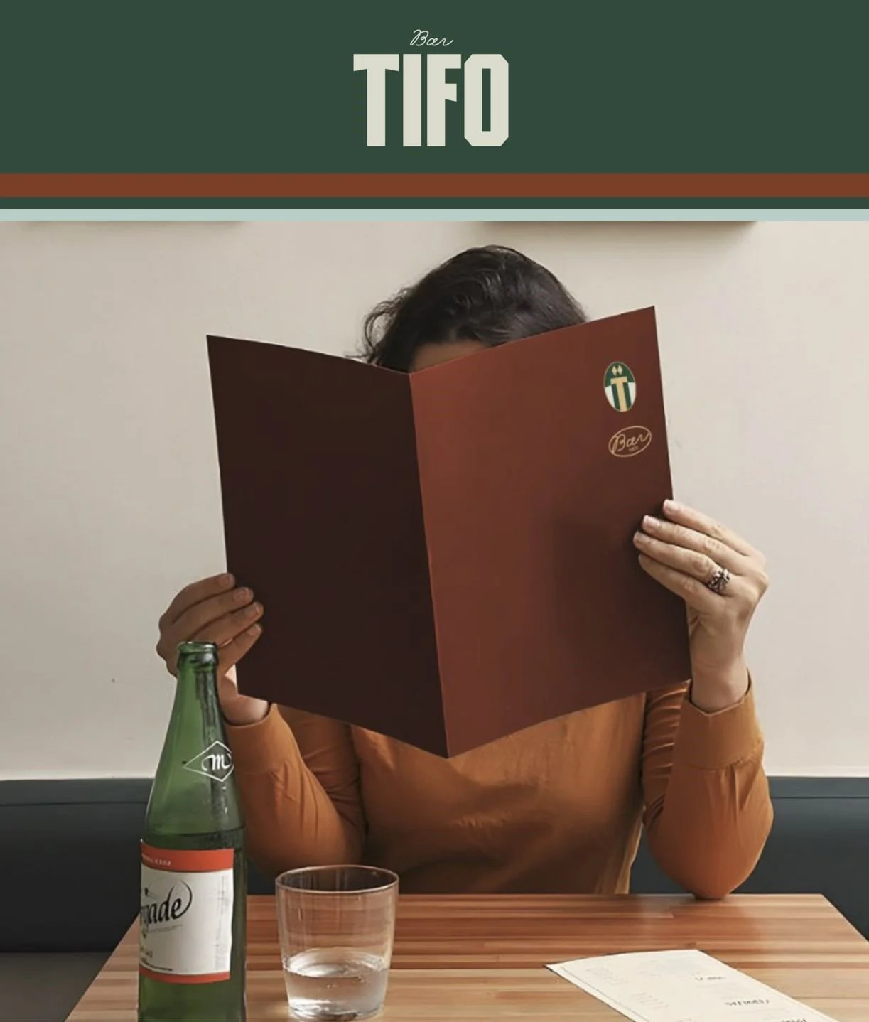 Person sits at a table in a restaurant named Tifo, holding a large menu that covers their face. The table has a bottle, a glass of water, and a menu sheet on it.