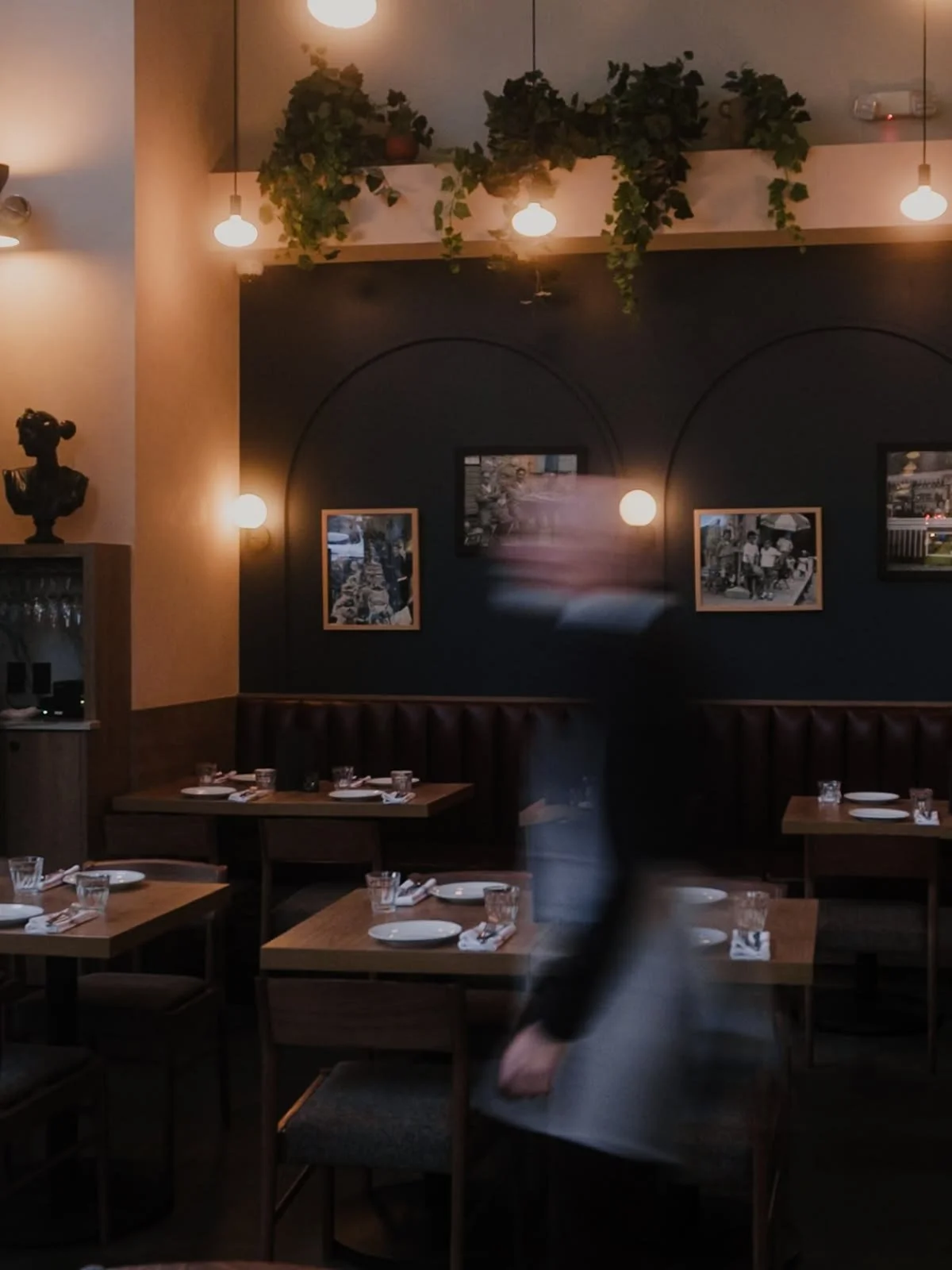 Interior of a restaurant with dim lighting, tables set with plates, glasses, and utensils, framed pictures on the navy blue wall, hanging pendant lights, a statue on a shelf, and some greenery on the ceiling.