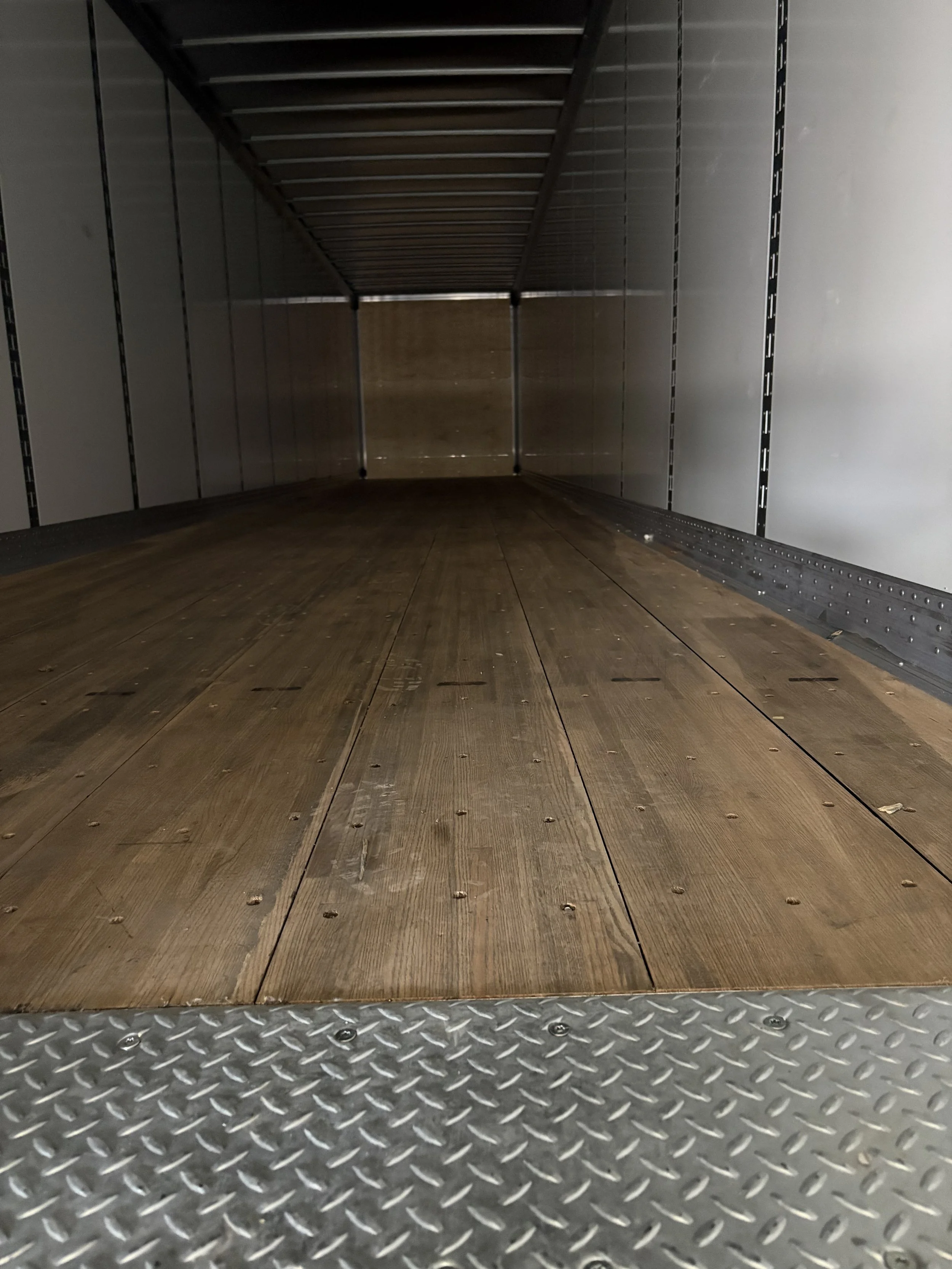 Looking inside an empty truck cargo area with wooden floorboards and silver walls, viewed from the open back door.