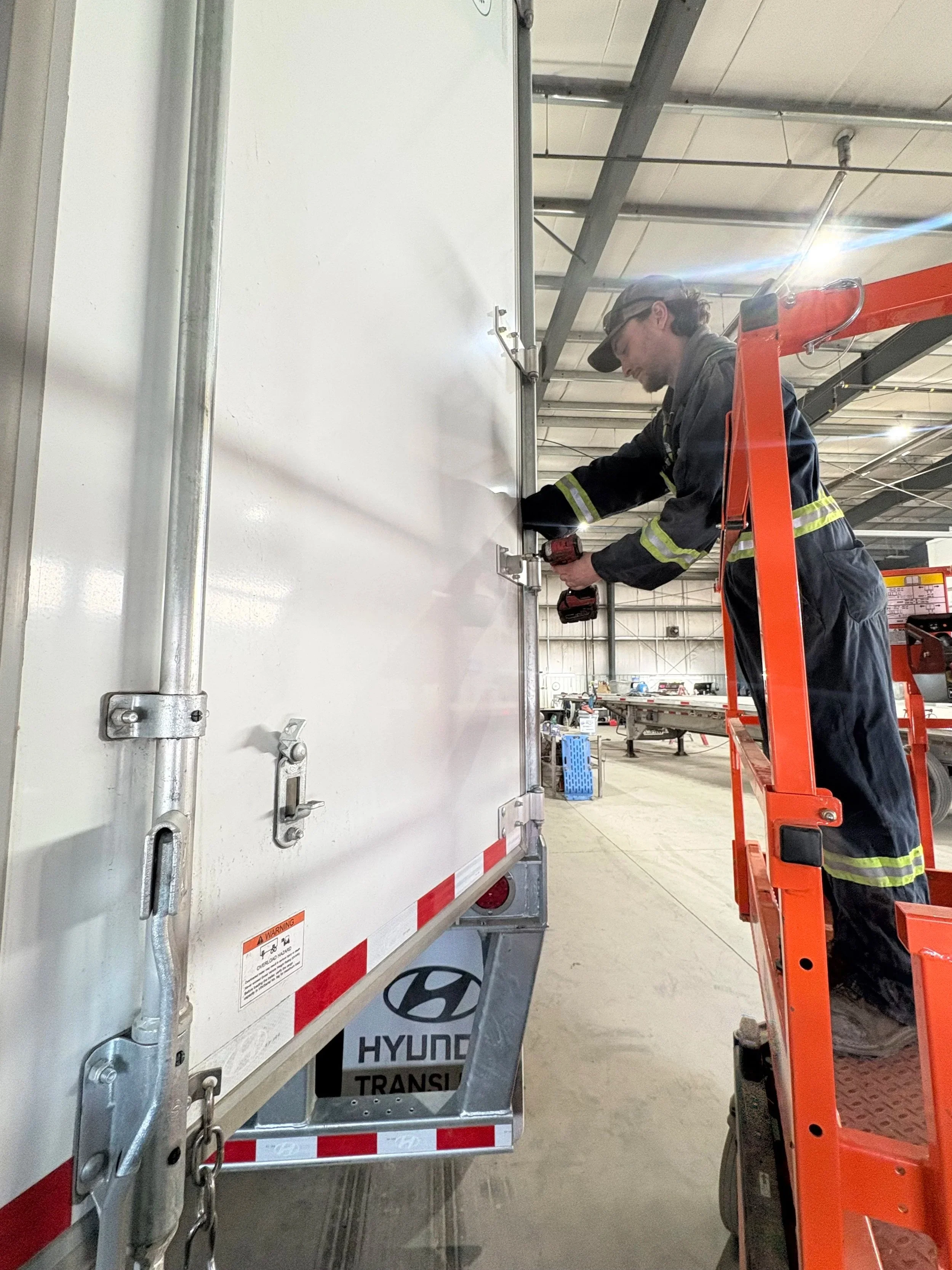 A man in safety gear and helmet installing or repairing a white truck container using a power drill, standing on an orange lifting platform inside a warehouse.