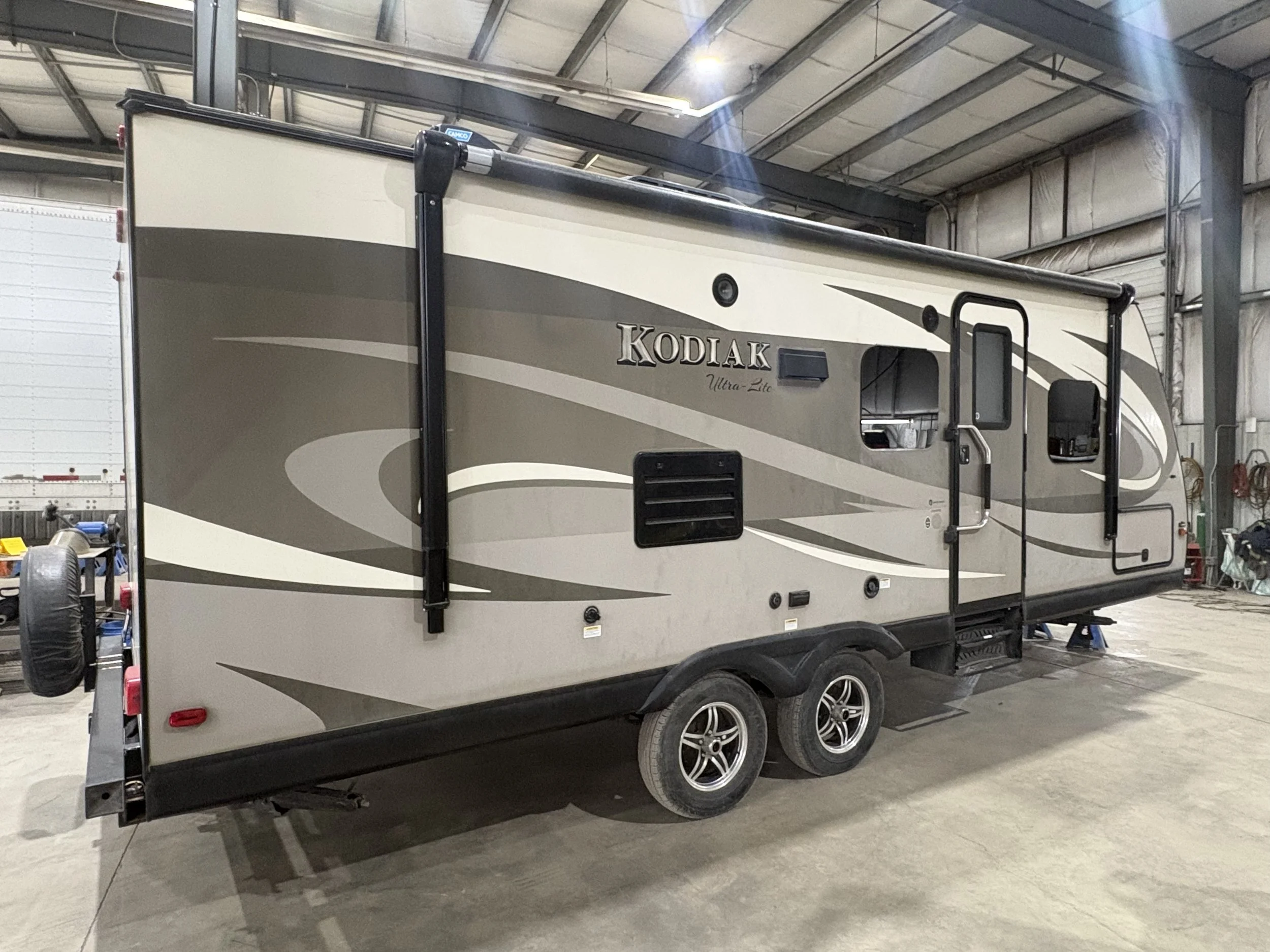 A Kodiak Ultra Lite travel trailer inside a warehouse with a high ceiling, metal beams, and a concrete floor. The trailer has a beige and brown exterior with black wheels, and there's a small window and door visible.