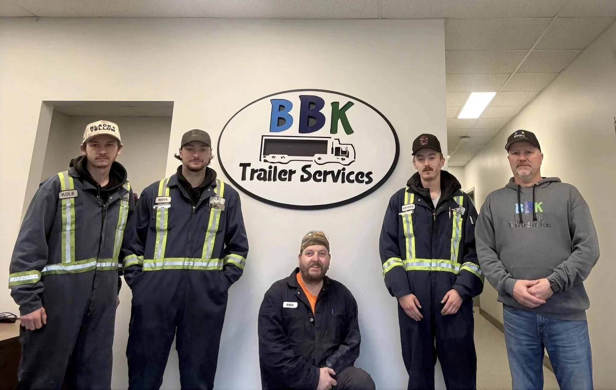 Five men in front of a BBK Trailer Services sign. The men are wearing coveralls and some are wearing hats. They are standing indoors against a wall with the BBK Trailer Services logo.