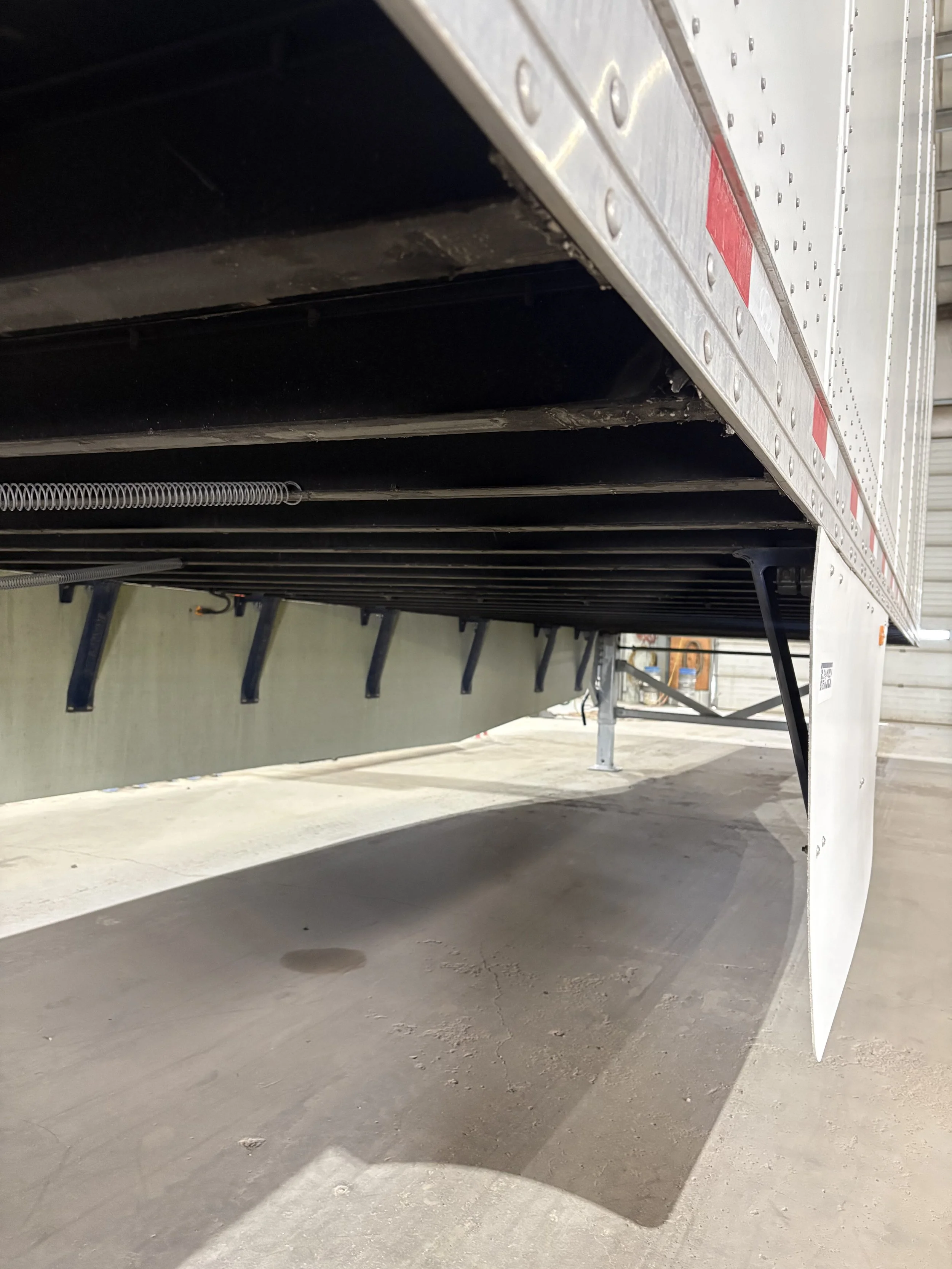 Side view of a large white semi-truck trailer parked on concrete ground, showing the underside of the trailer and support legs.