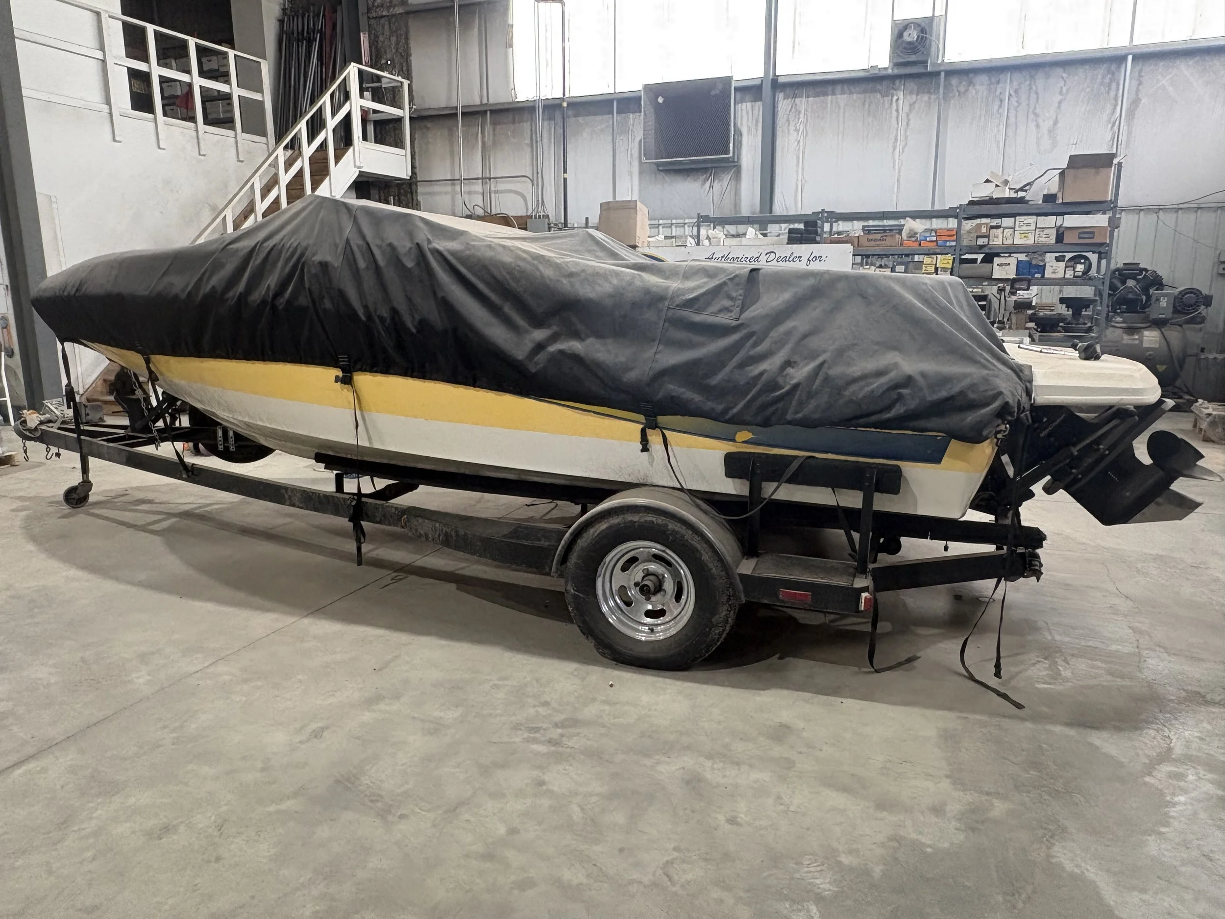 A boat on a trailer inside a warehouse with metal shelves, boxes, tools, and equipment in the background.
