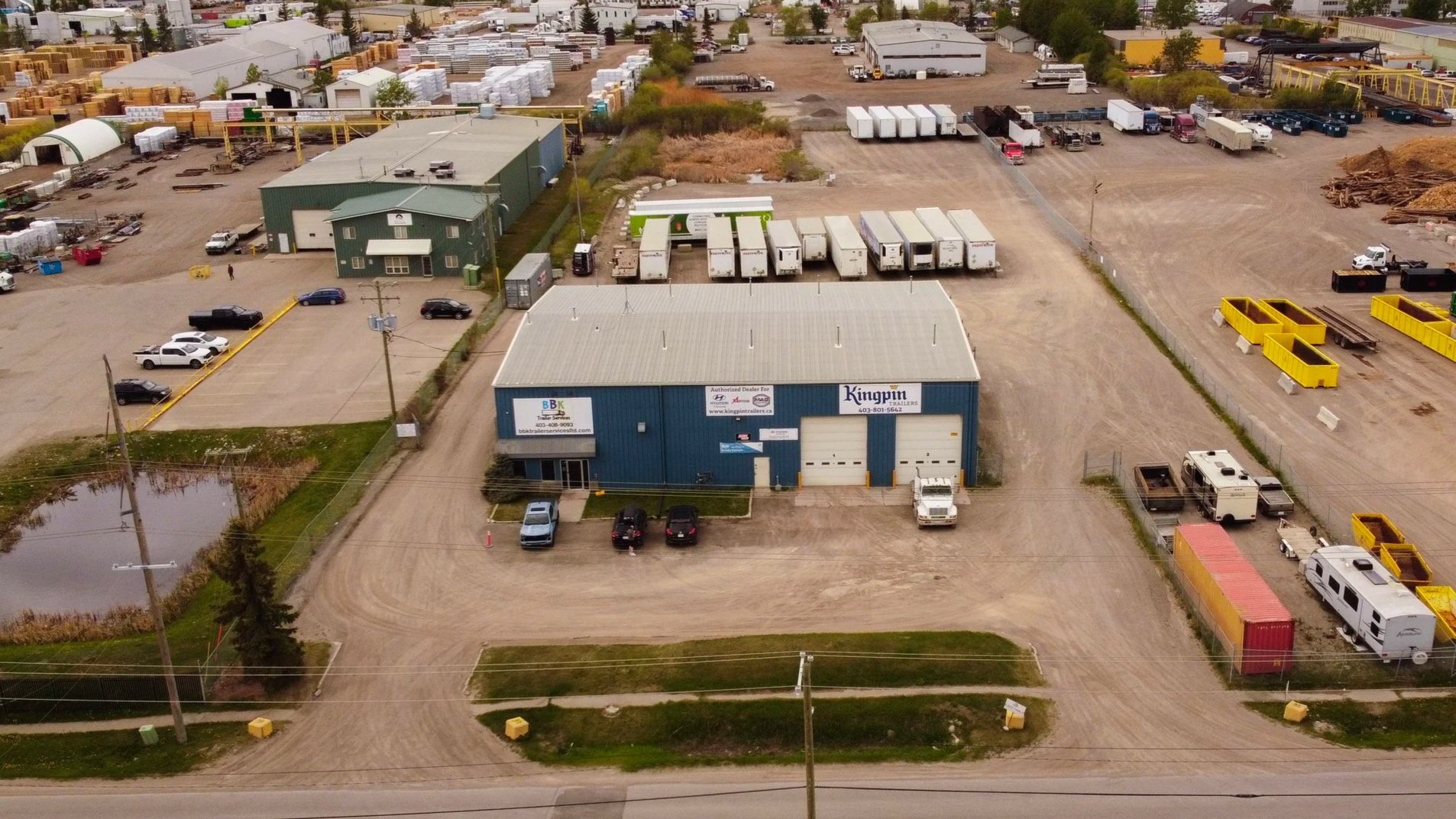 An aerial view of an industrial trailer sales lot showing trucks, cars, storage units, and a large blue and gray building with signage.