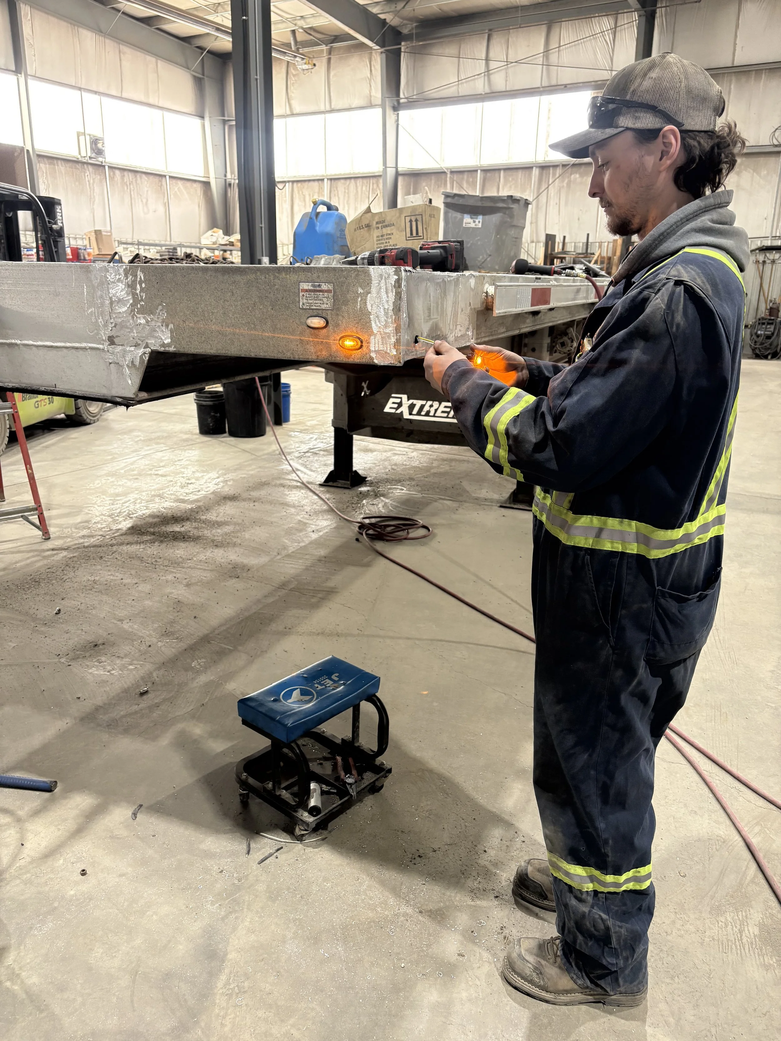 A worker in safety gear welding a large metal structure inside an industrial warehouse. There are tools and equipment nearby, and the worker is focused on the task.