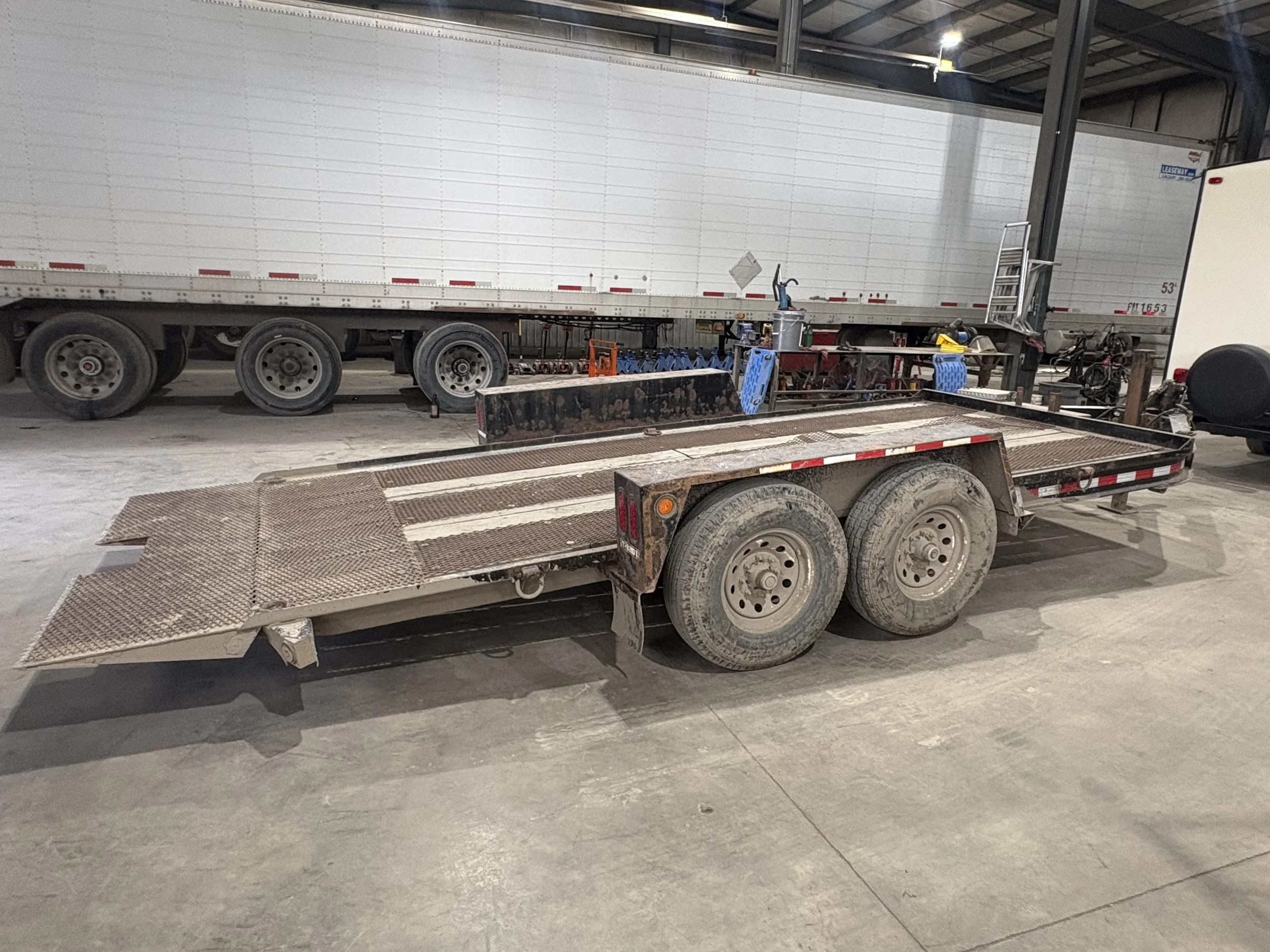 A white semi-truck parked inside a large warehouse or garage, with a flatbed trailer attached. The surroundings include various tools and equipment, with bicycles in the background. The area is well-lit with overhead lighting.