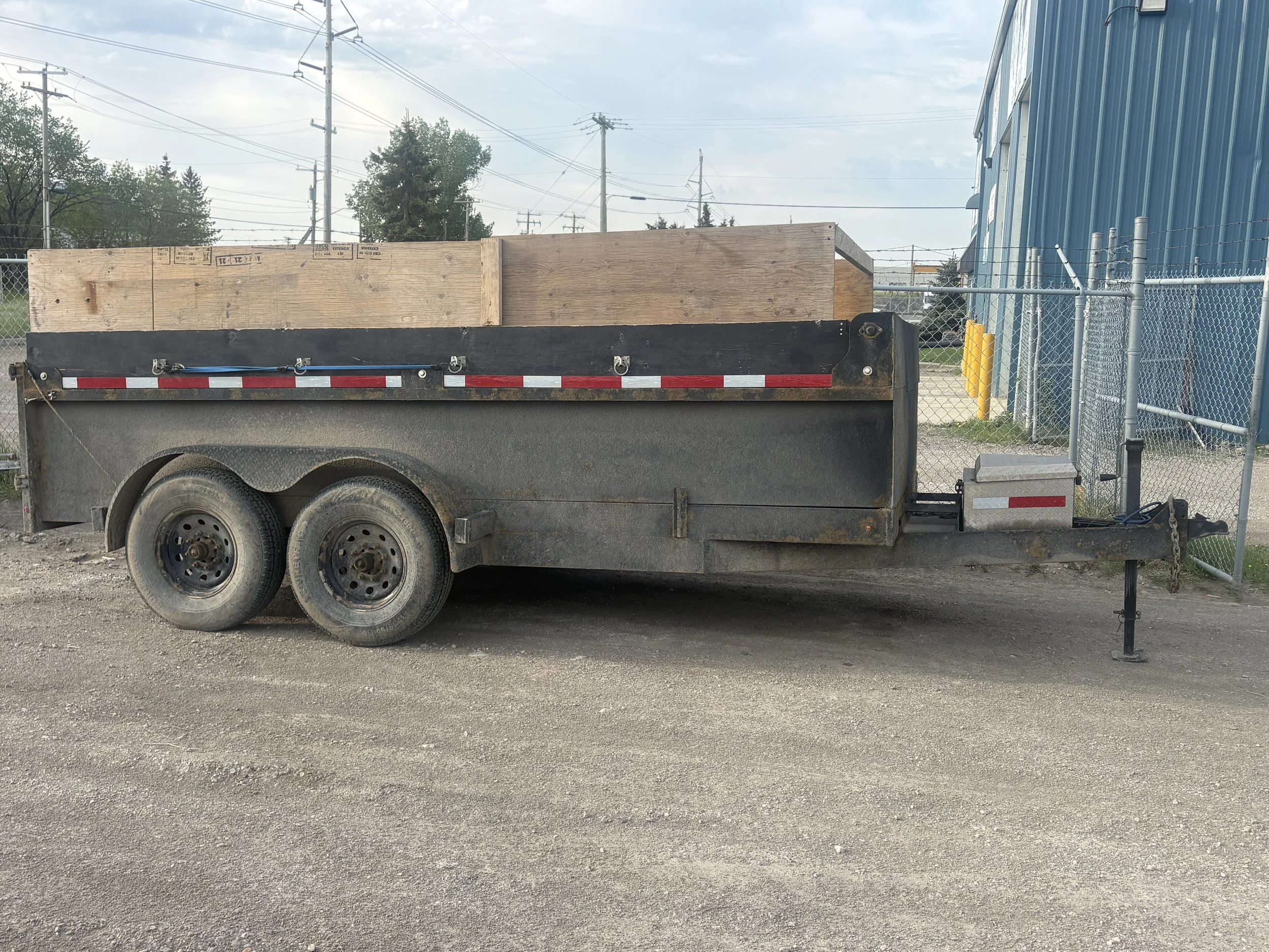 A flatbed utility trailer parked on a gravel lot, loaded with wooden planks, with a chain securing the load. In the background, there is a chainlink fence, industrial buildings, utility poles, and trees.