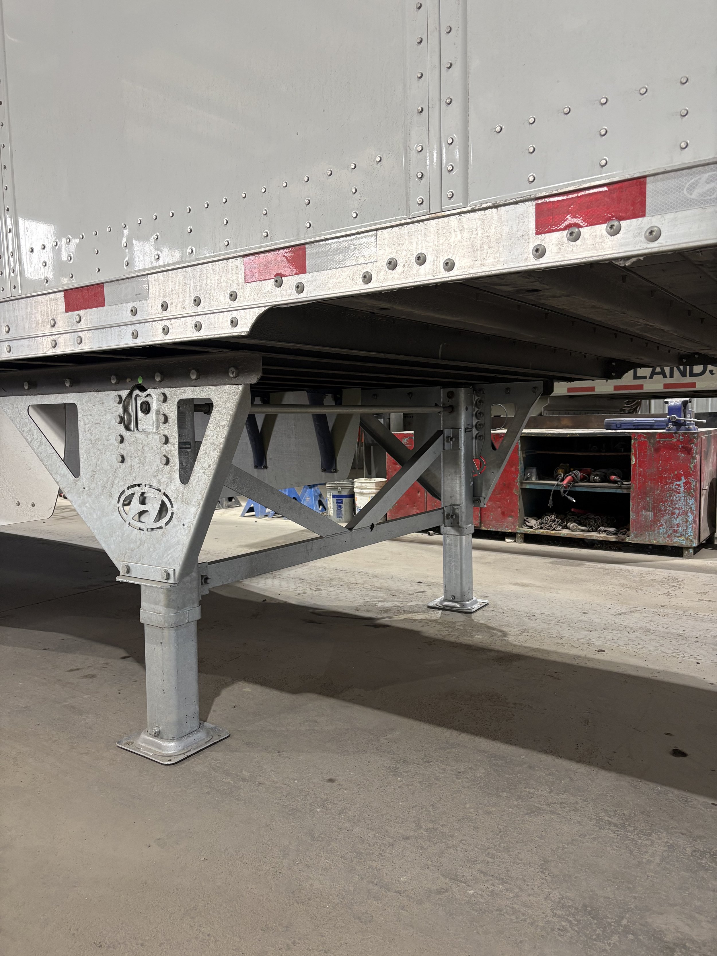 Close-up of a metal trailer or truck with structural support legs, reflective red and white safety striping, parked on concrete floor.