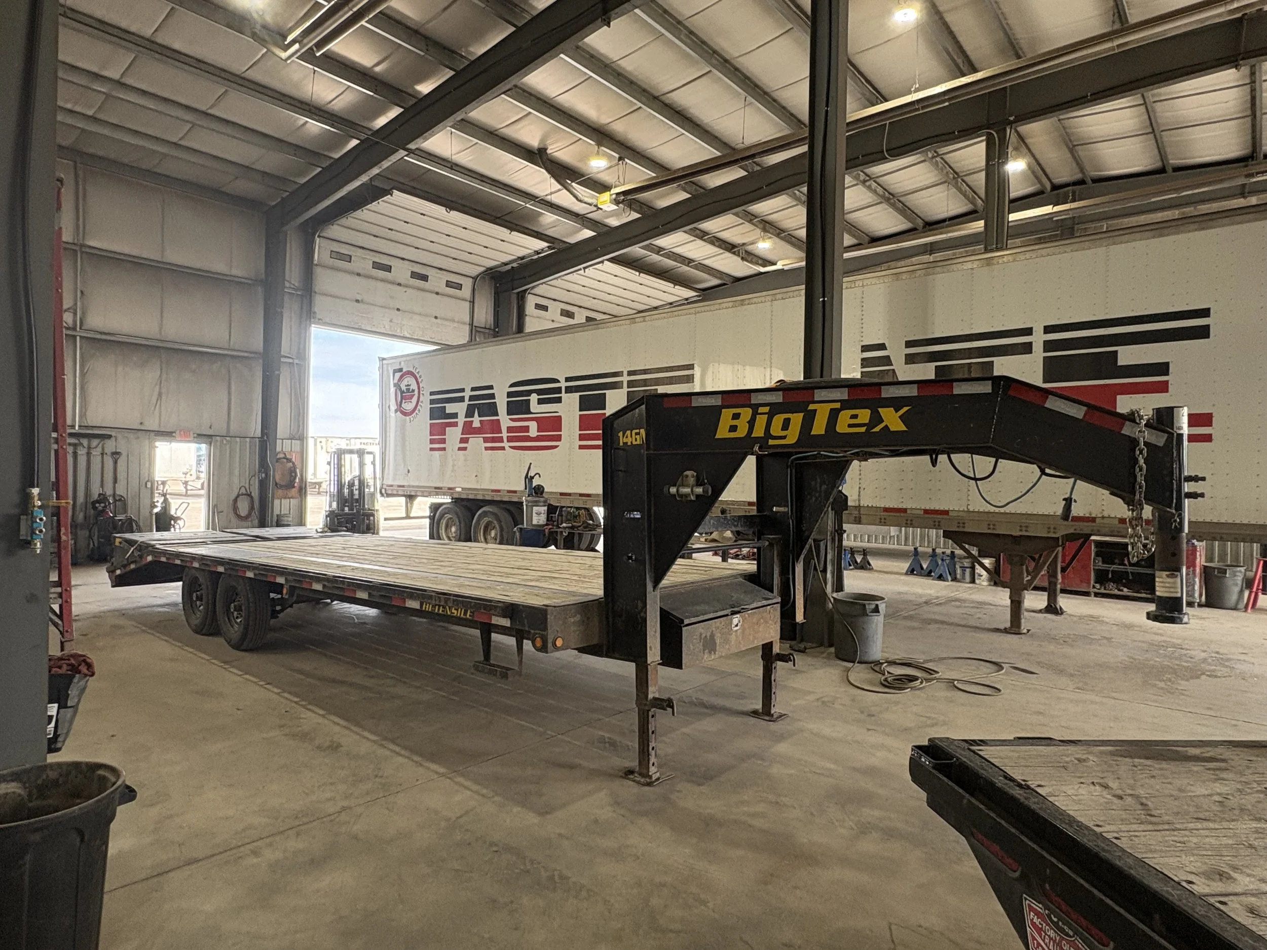 Interior of a warehouse or garage with a large truck labeled 'Fast' parked inside. There is equipment, a flatbed trailer, and tools visible in the space.