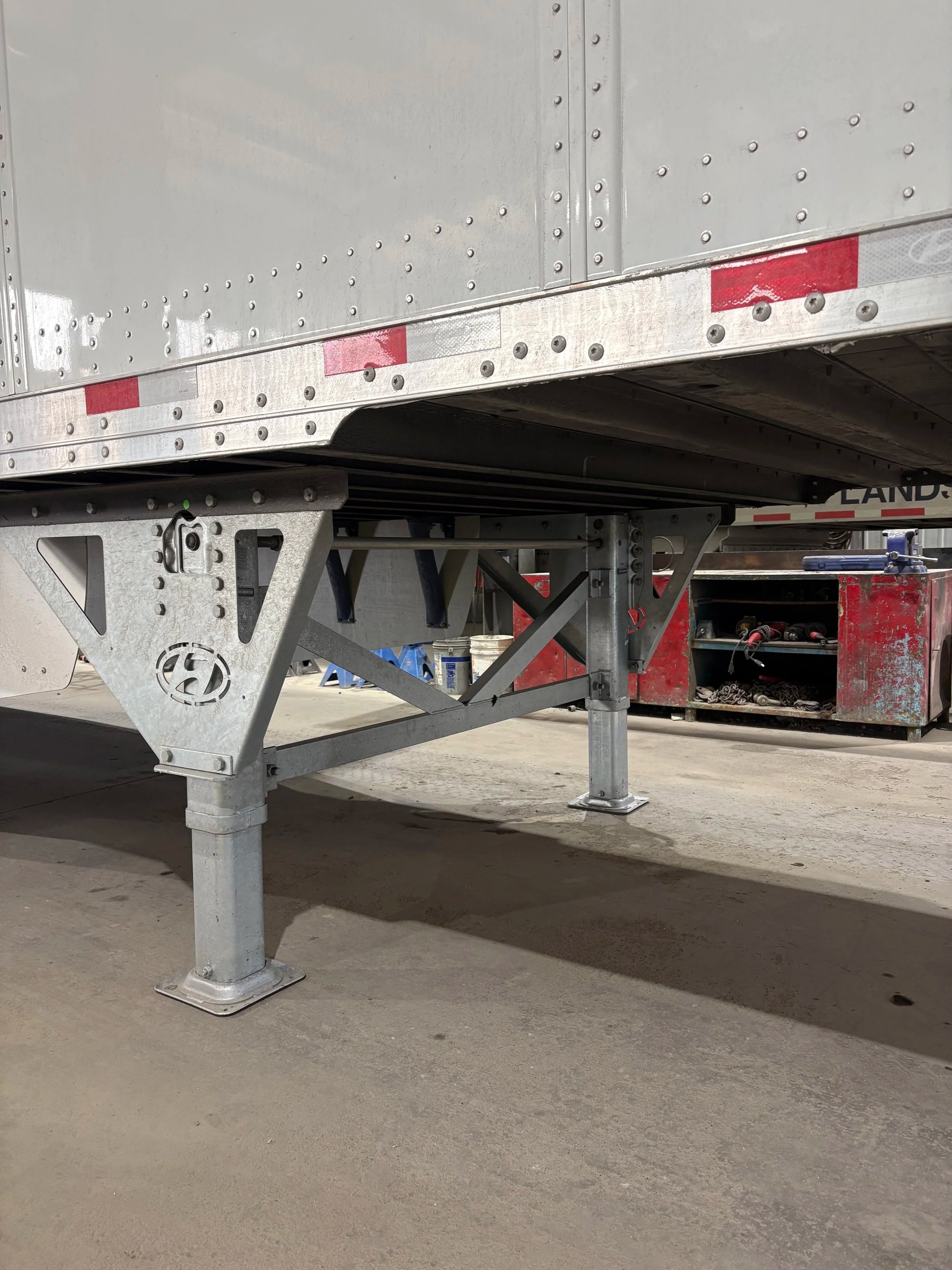 Close-up of the underside of a silver trailer with a metal support stand on a concrete floor.
