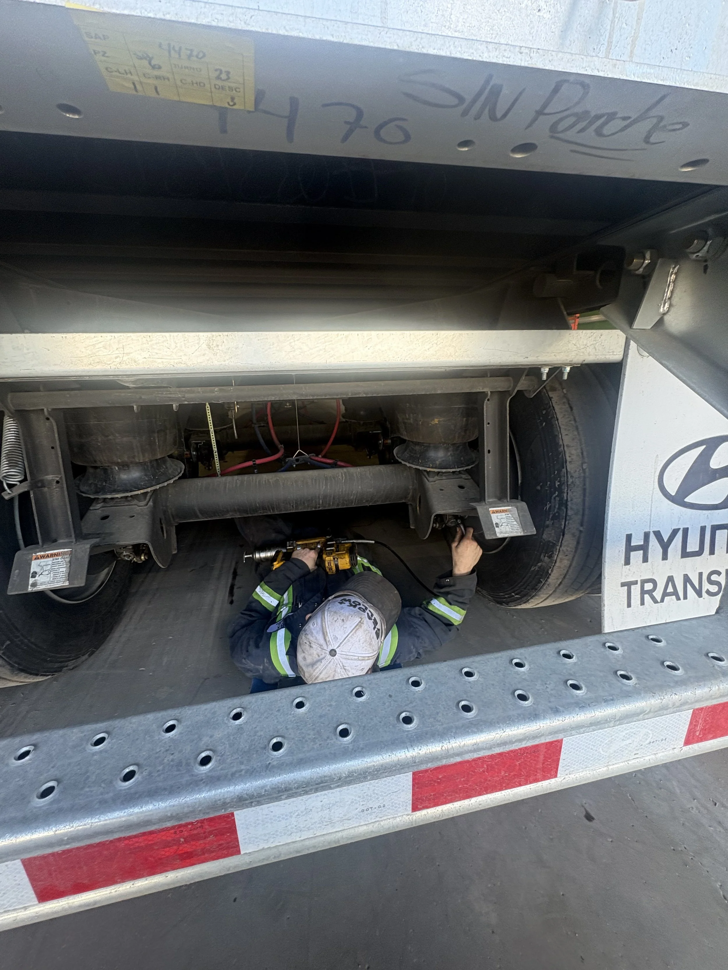 A worker wearing a helmet and safety vest working under a large vehicle, possibly a truck, inspecting or repairing components underneath.