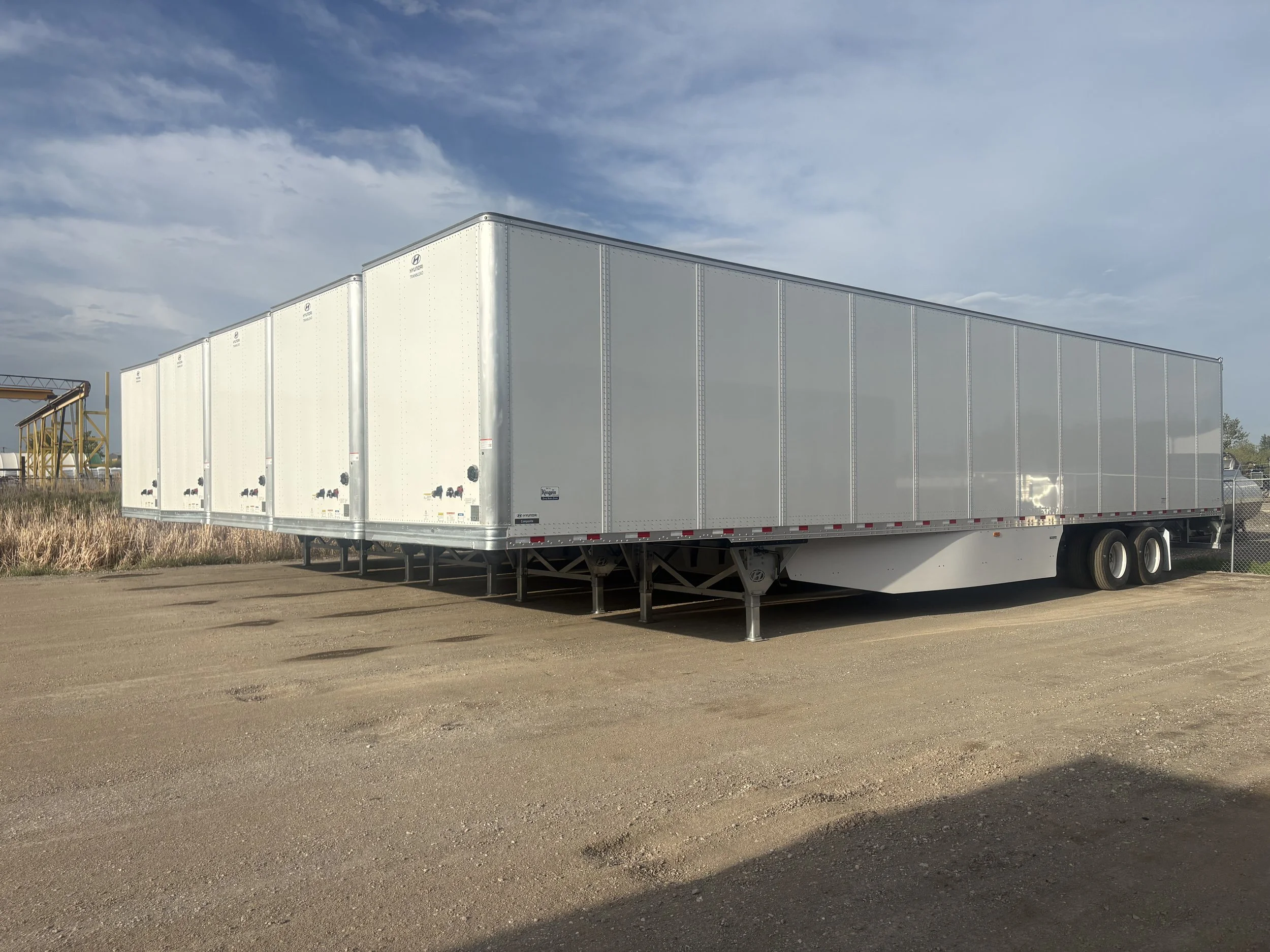 A white semi-truck trailer parked on a gravel lot under a partly cloudy sky.