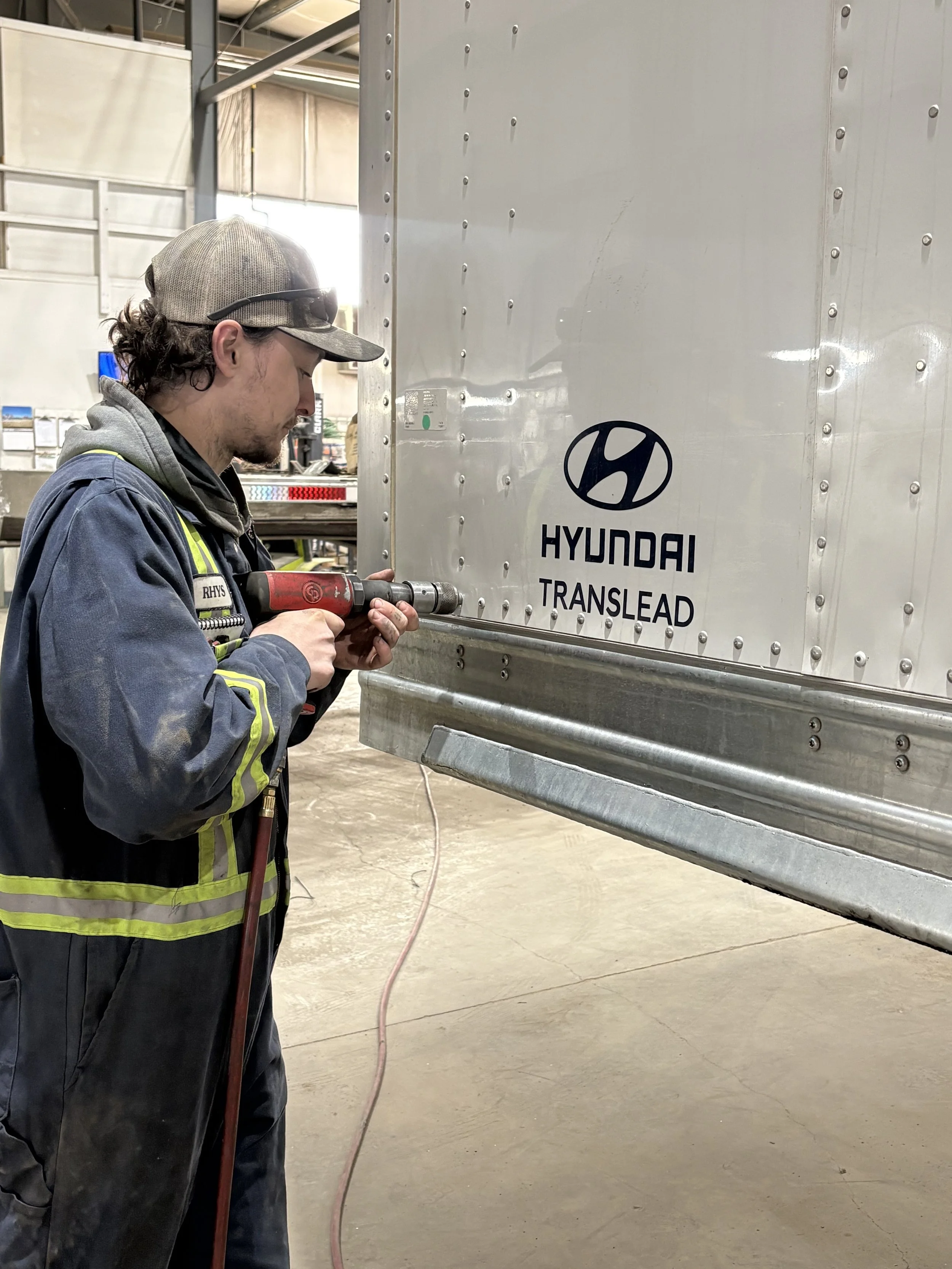 A man working in a warehouse or garage, using a power drill on a large white metal surface with Hyundai Translead branding. He wears a cap, safety glasses, and a jacket with reflective stripes.