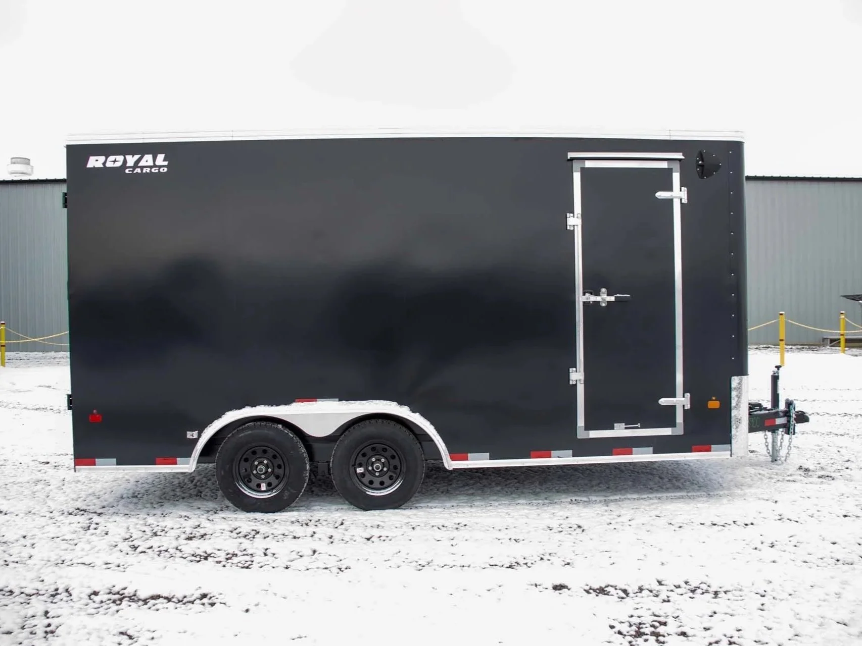 Black cargo trailer with a roll-up door, sitting on snowy ground outside near a gray industrial building.