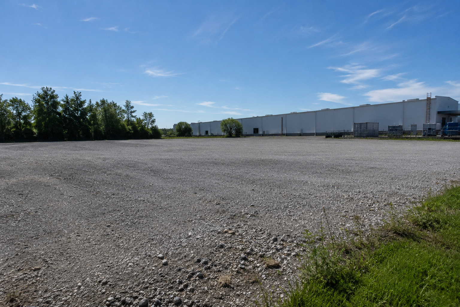 Empty gravel lot with trees and a large white industrial building under a blue sky with sparse clouds.
