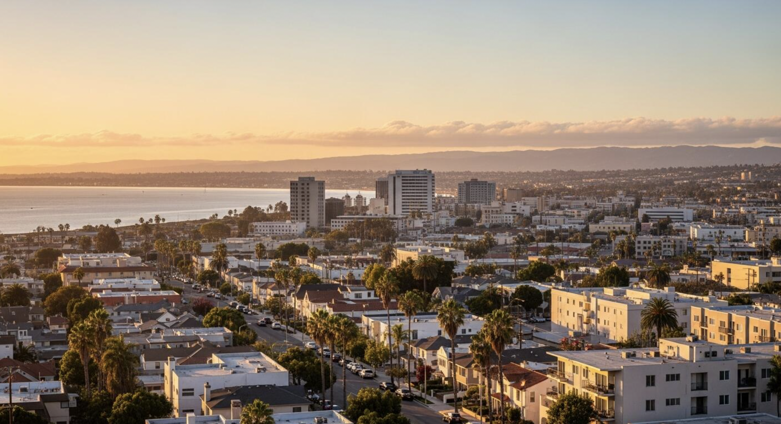 Sunset over a cityscape with high-rise buildings, residential houses, palm trees, and a body of water with mountains in the background.