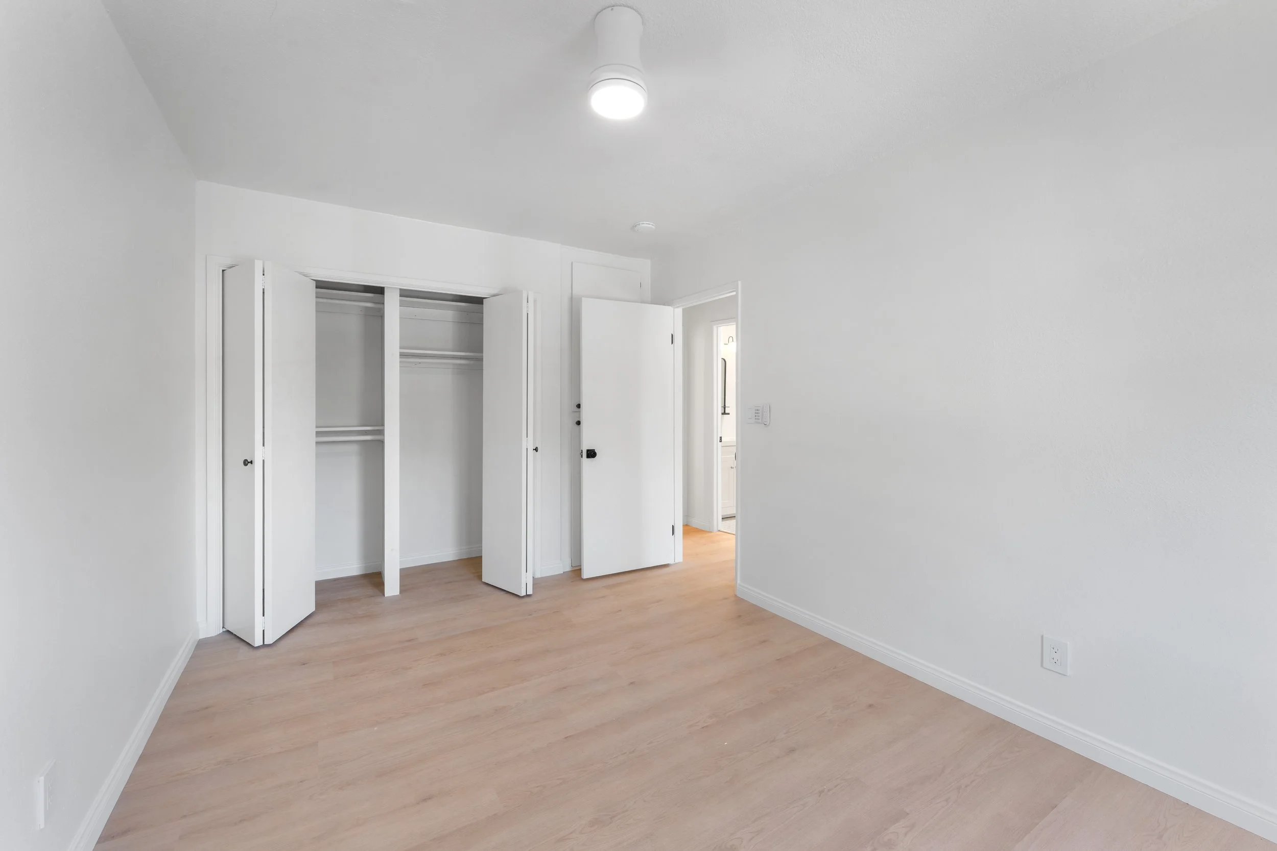 Empty room with white walls, hardwood floors, a closet with open bi-fold doors, and a doorway leading to another room.