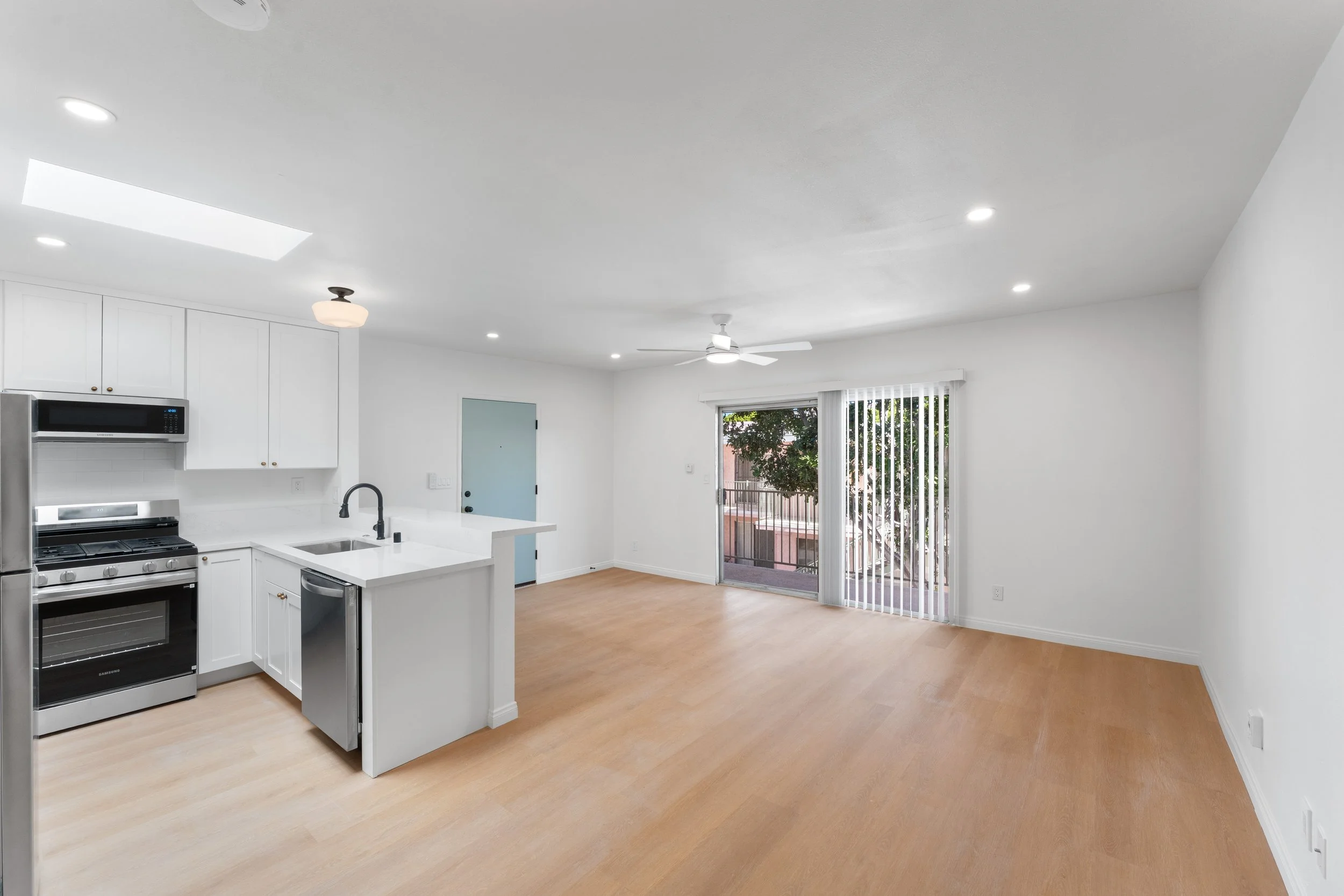 Empty living area with kitchen, sliding glass door, and ceiling fan in a modern apartment.