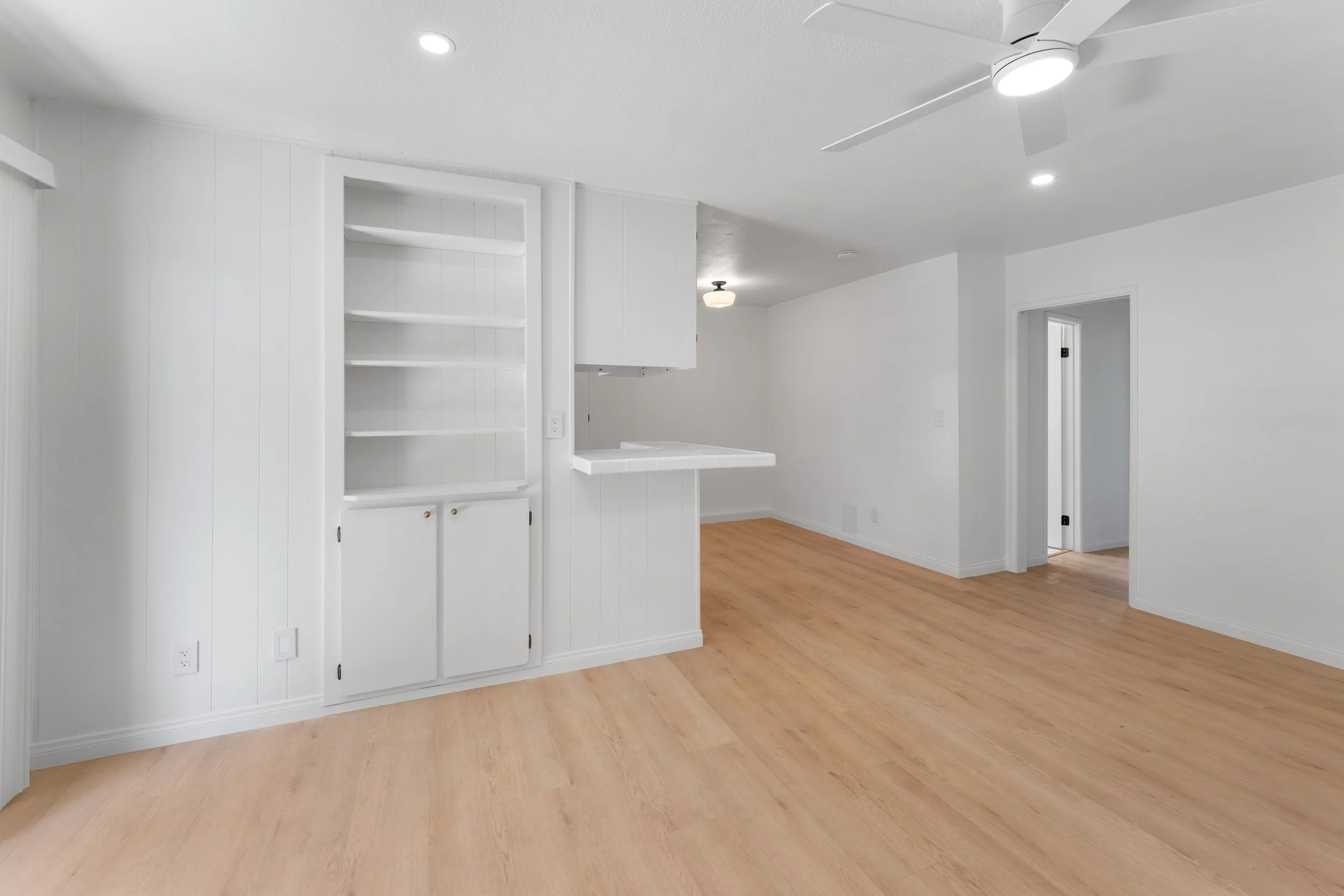 Empty room with white walls and ceiling, wooden floor, built-in white shelving, and ceiling fan with light.
