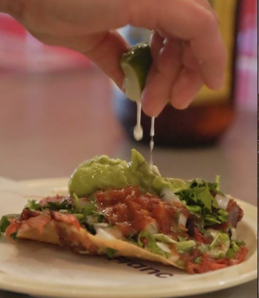 A hand squeezing lime onto a slice of pizza topped with tomatoes, onions, and greens.