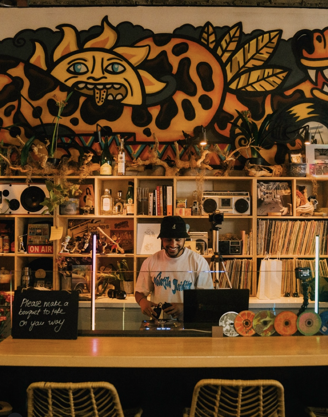 A man smiling behind a DJ setup in a colorful, vibrant room with a large graffiti mural of a dragon and a moon face on the wall. The room is decorated with books, plants, and vinyl records, giving an artistic and lively atmosphere.
