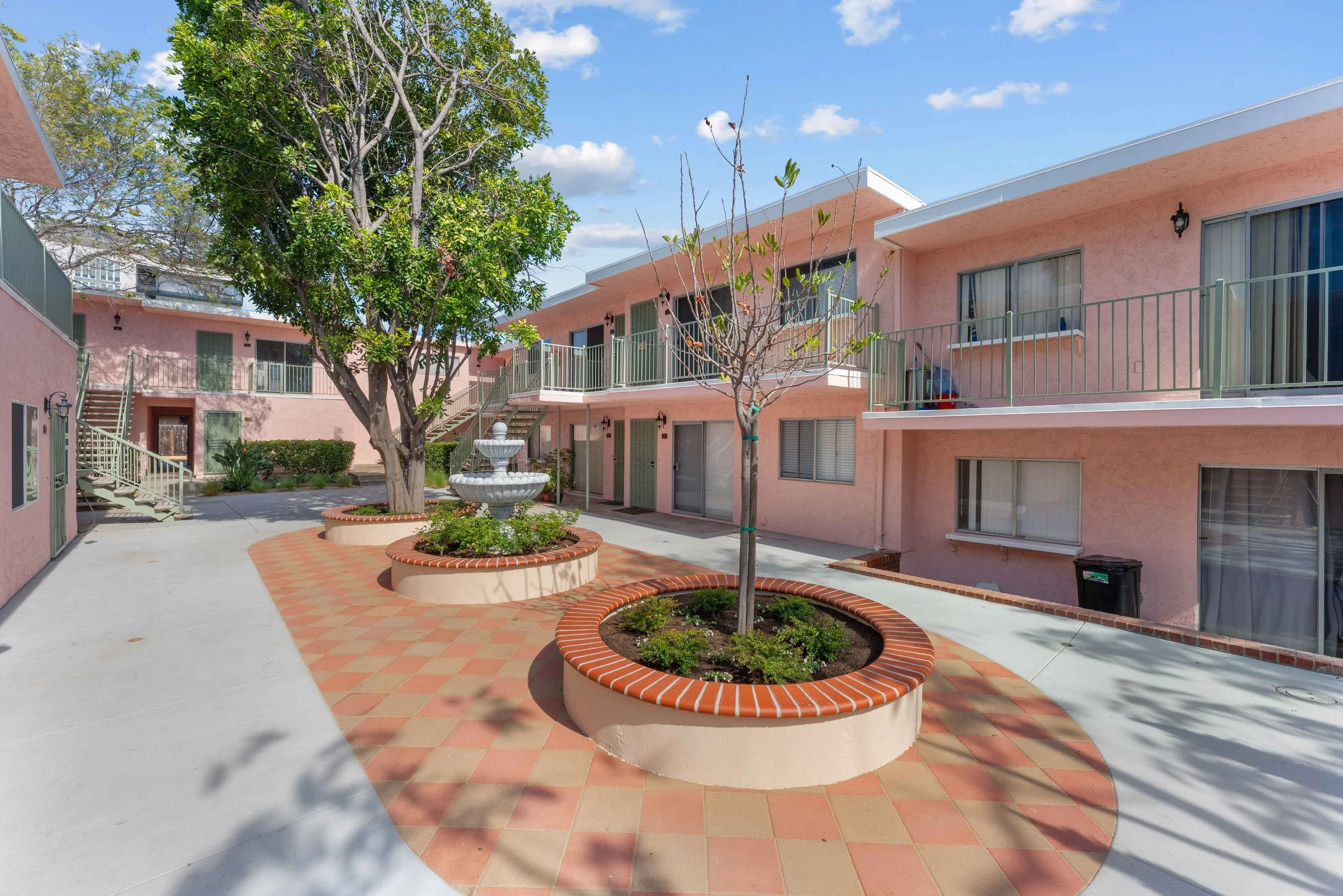 Courtyard of a pink apartment building with green railings, two trees, a fountain, and brick-edged planter circles, under a blue sky with clouds.
