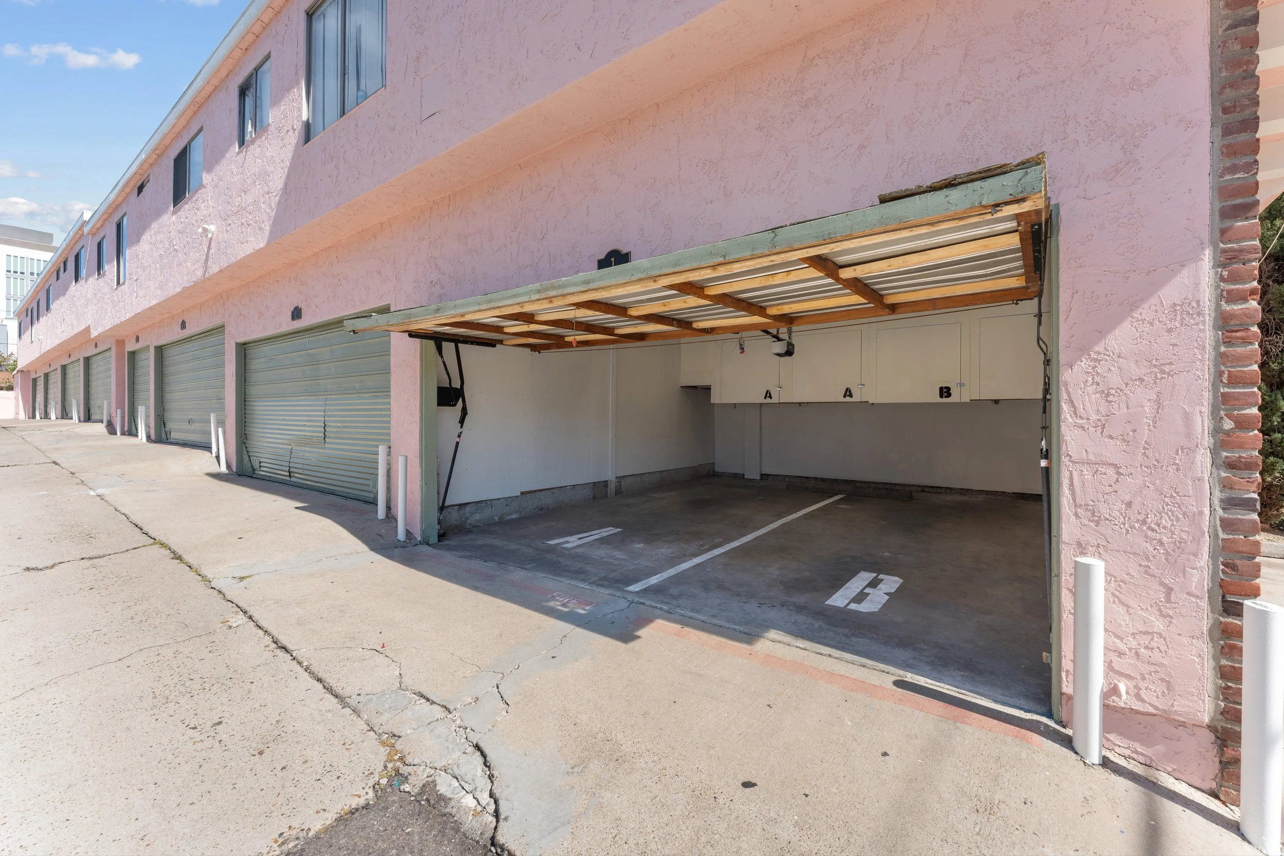 A parking garage with pink exterior walls and a row of closed silver garage doors. One parking space is visible, marked with a white number 3, under a makeshift wooden and metal roof extension. The pavement in front shows cracks.