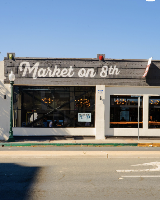 Exterior view of a restaurant called 'Market on 8th' with large windows, sidewalk, and street in front, under a blue sky.