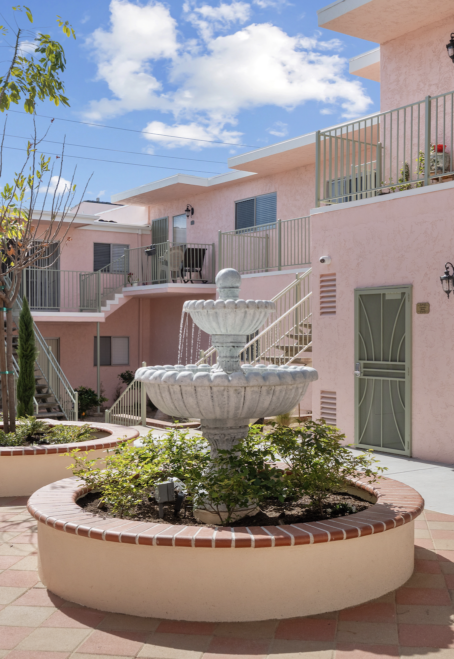 A pink apartment building with outdoor balconies, metal railings, and stairs. A white stone fountain with water flowing into a garden bed with small plants in the courtyard. Paved walkway, trees, and blue sky with clouds.