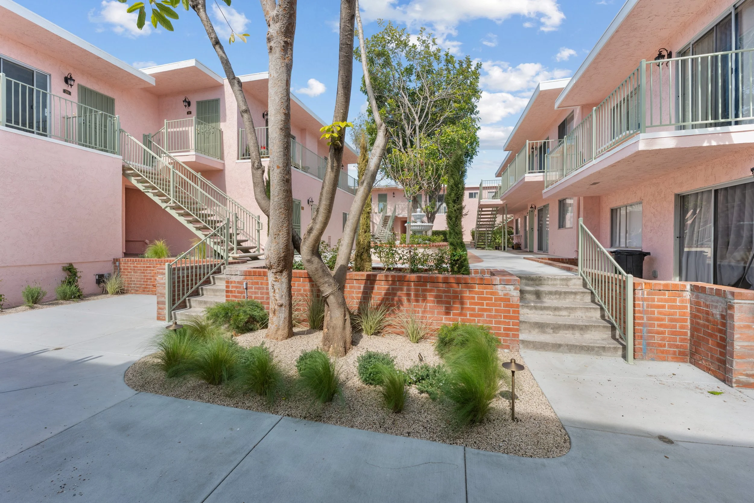 Apartment courtyard with pink buildings, exterior stairs, trees, and a fountain under a partly cloudy blue sky.