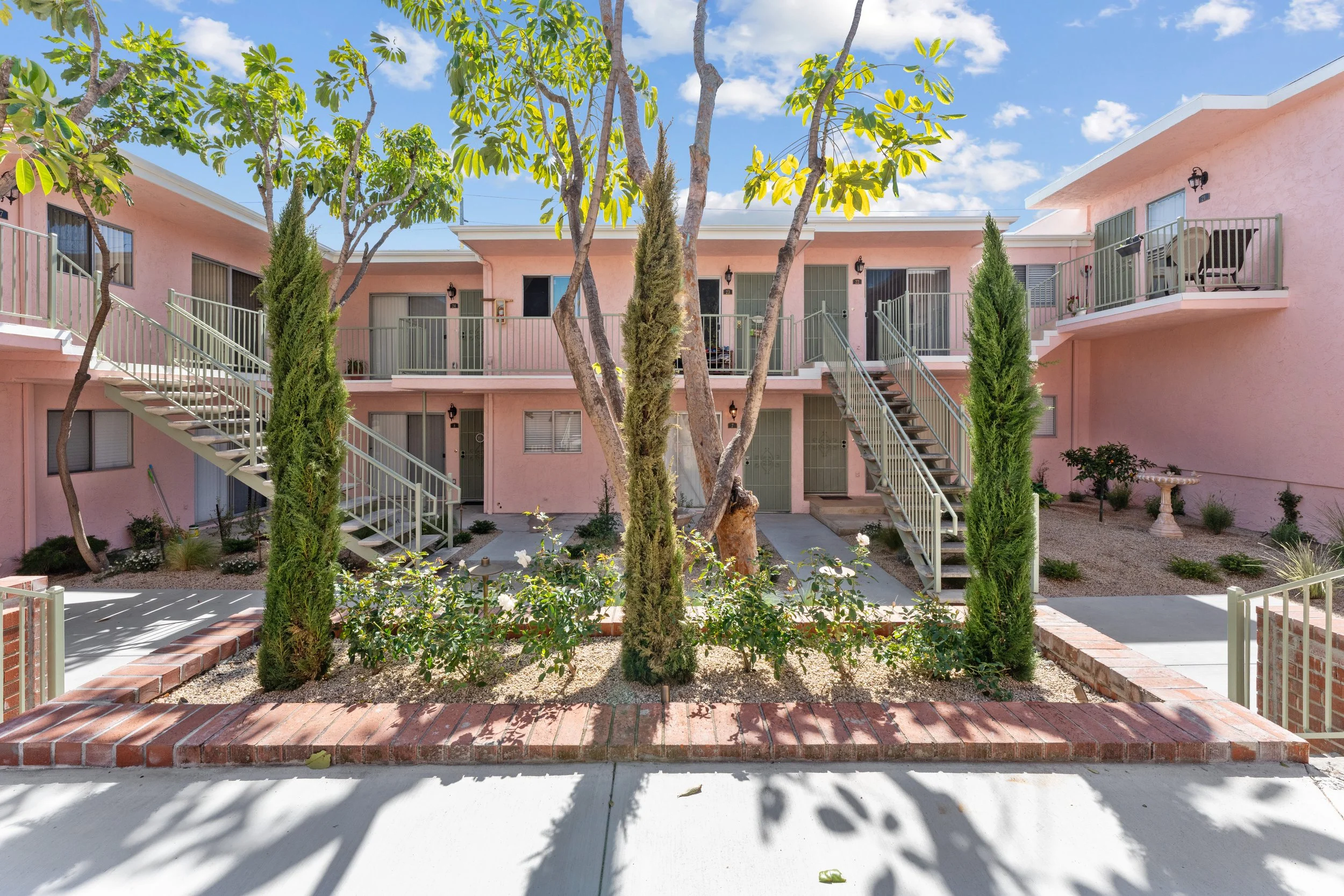 The image shows a pink apartment building with stairs leading to the upper units, surrounded by trees and small landscaped areas with plants and bushes. There are balconies with outdoor furniture and a bright blue sky with some clouds.