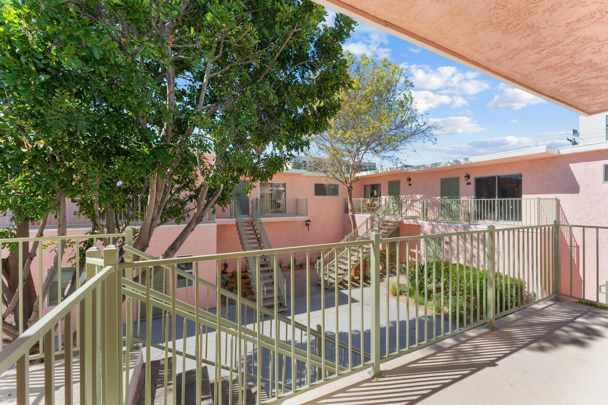 A view of a pink residential building complex with outdoor stairs, trees, and a yellow fence, under a partly cloudy sky.