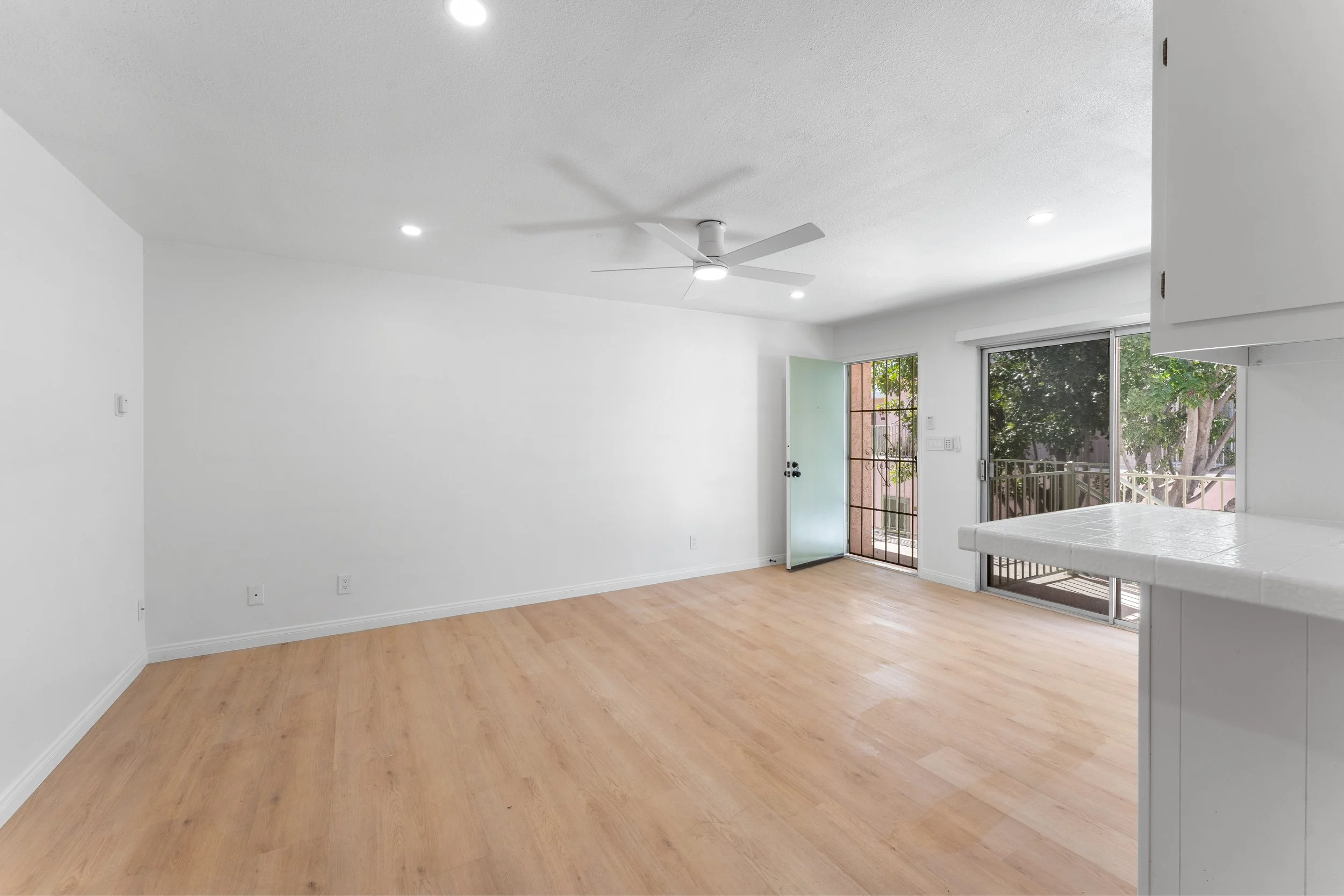 Empty living room with light wood flooring, white walls, ceiling fan, recessed lighting, sliding glass door, and partially visible kitchen countertop.