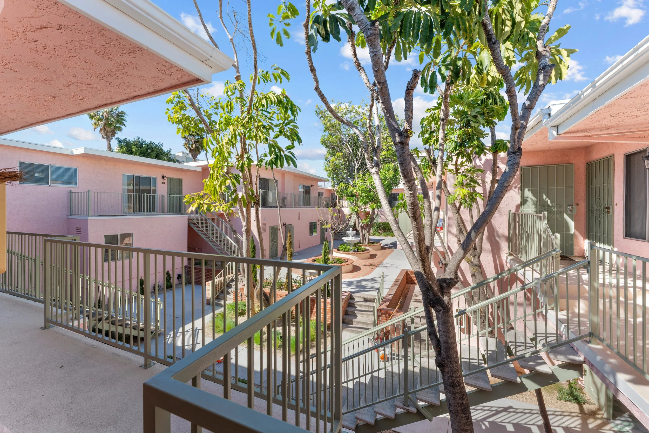 View of a pink apartment complex with metal railings, multiple levels, staircases, trees, and a fountain in the courtyard under a blue sky with scattered clouds.