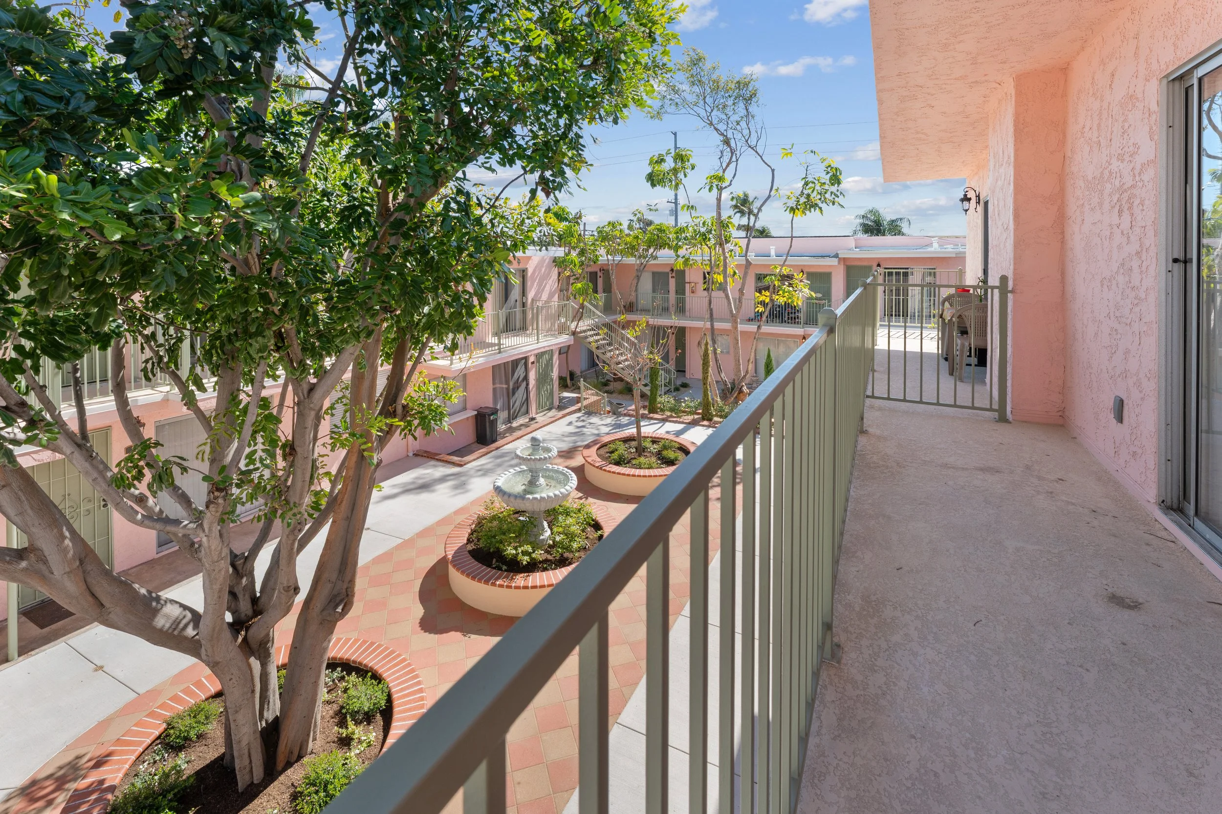 View from a balcony overlooking a courtyard with trees, potted plants, pink apartment buildings, and a fountain, under a blue sky with some clouds.