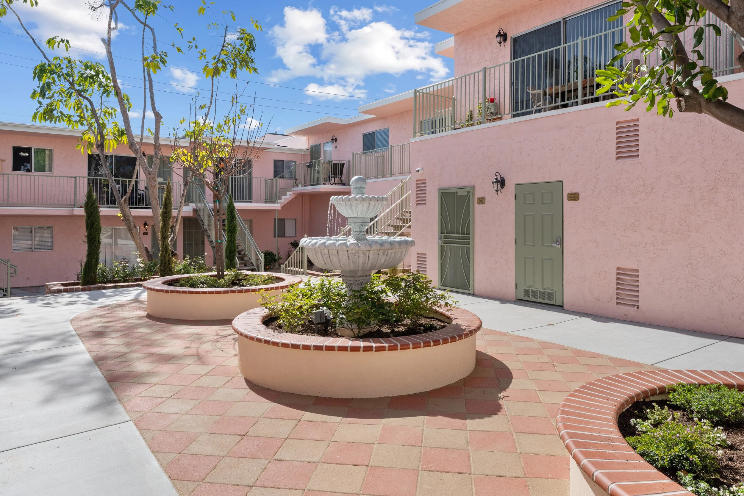 Outdoor courtyard with pink buildings, a white fountain, and planted trees under a blue sky with clouds.