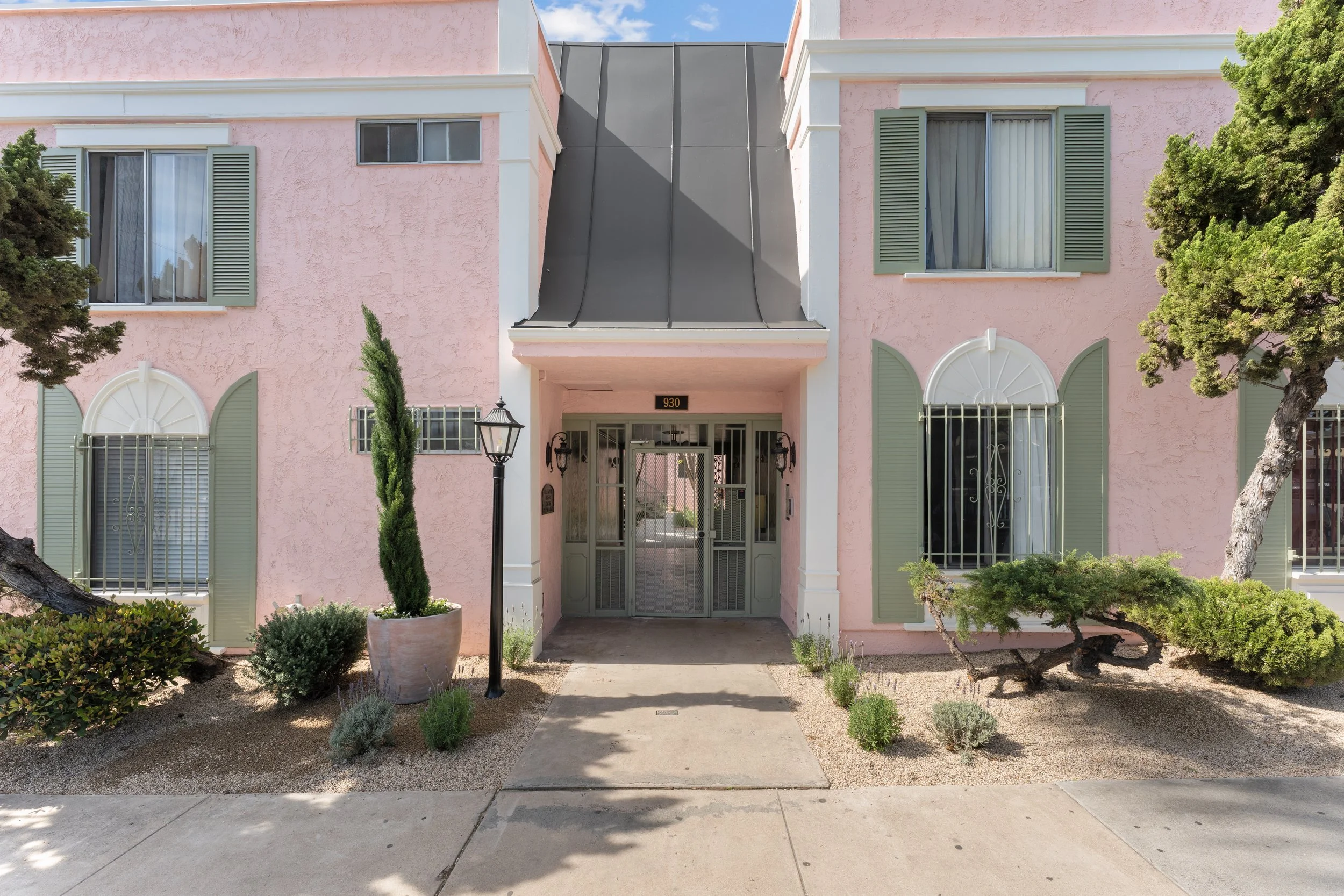 Front view of a pink residential building with green shutters, a gated entrance, and landscaped bushes and trees, under a partly cloudy sky.