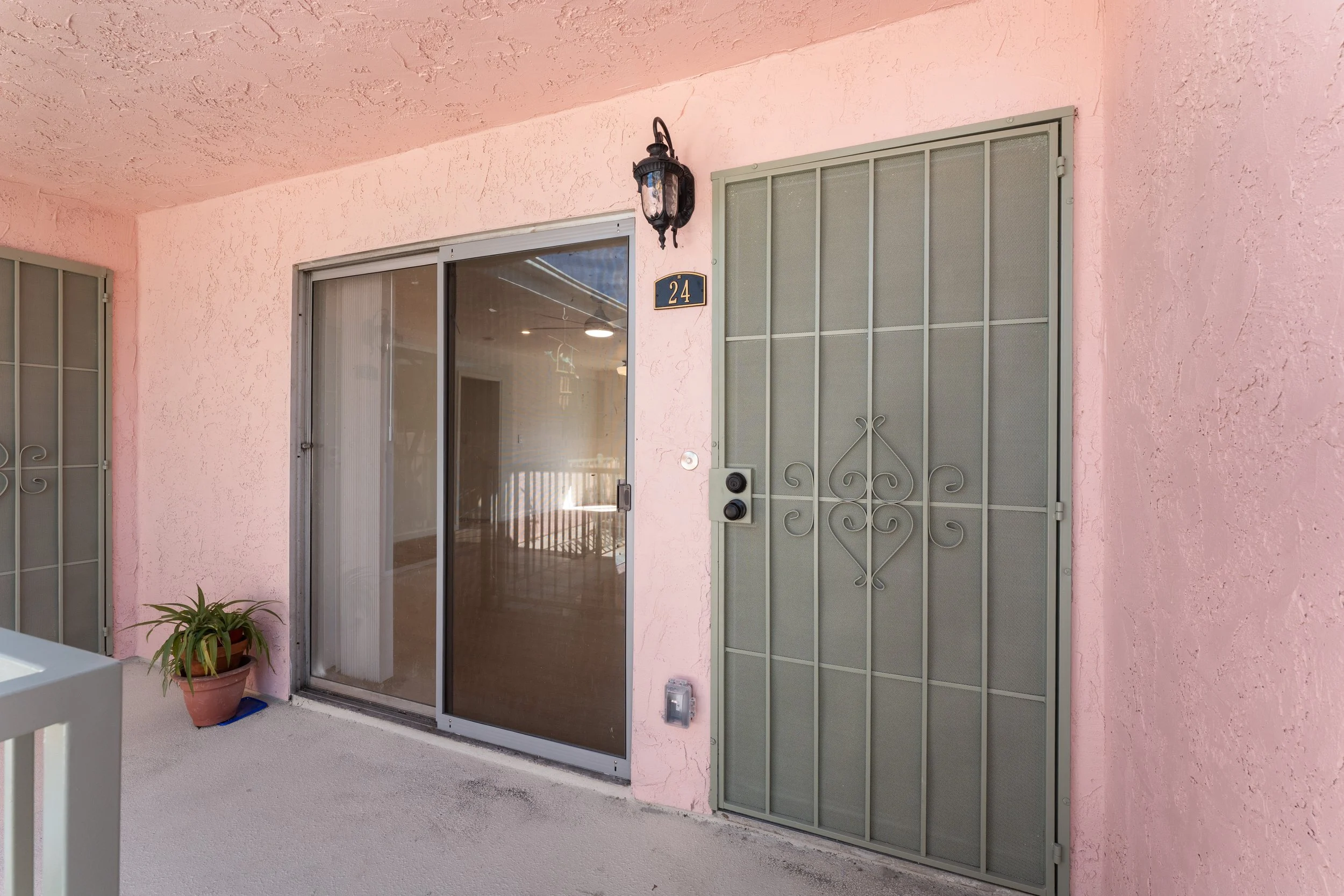 Exterior view of a pink stucco apartment balcony with sliding glass door, house number 24, and a metal security door, with a potted plant in the corner.