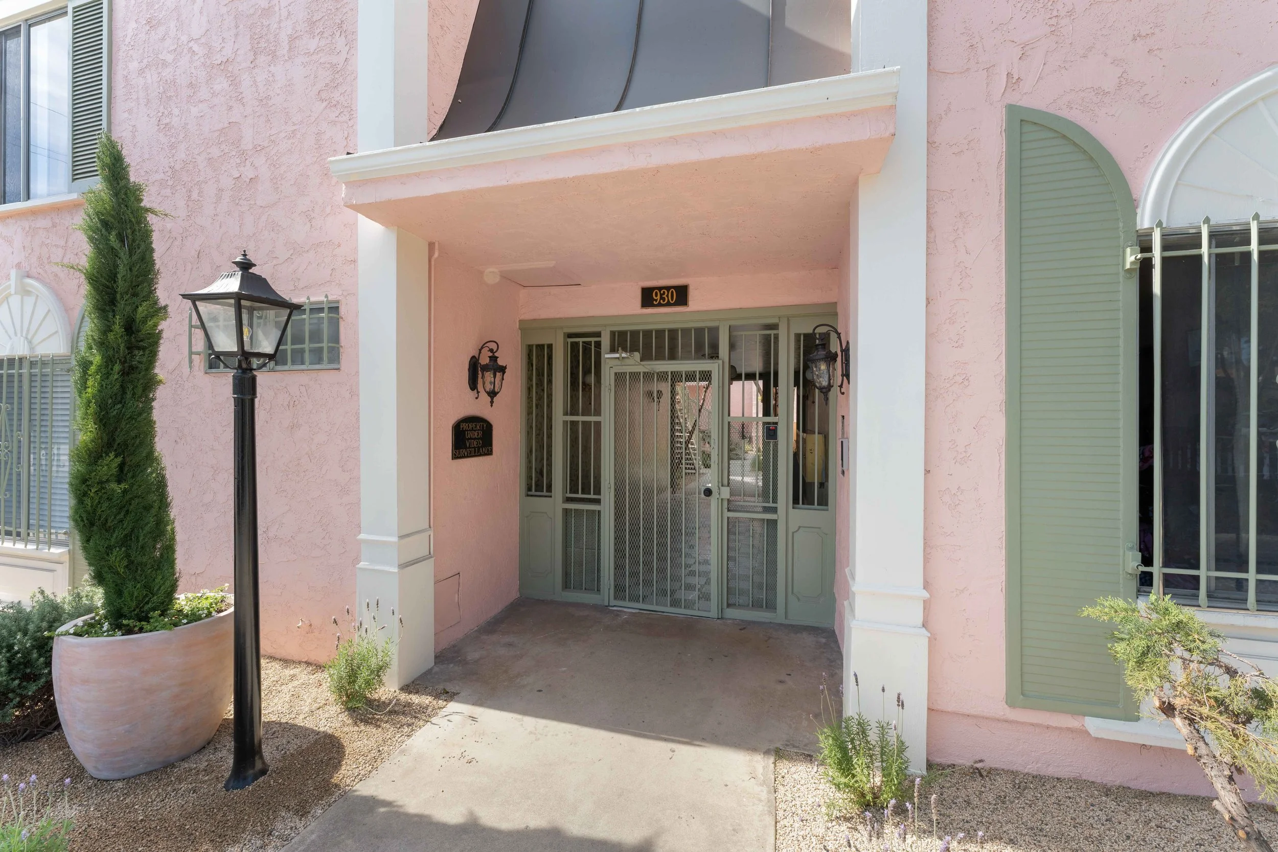 Pink residential apartment building entrance with green gate, black vintage-style lamppost, small bushes, and potted plant.