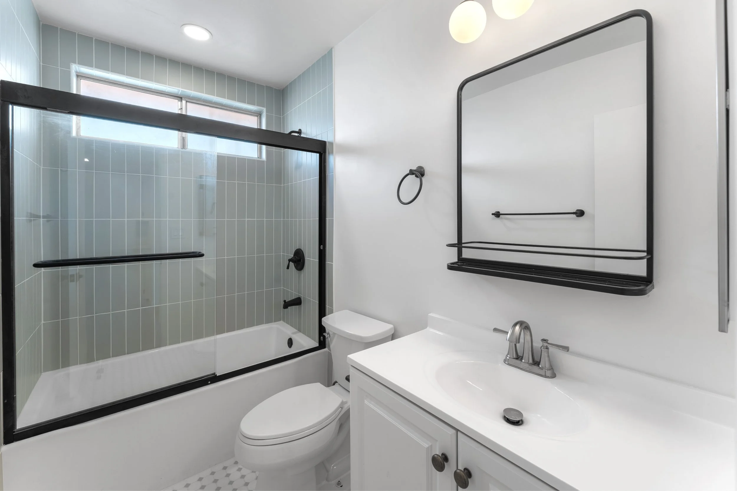 A modern bathroom with a glass-enclosed shower, a white toilet, and a white vanity with a sink and above-mirror storage. The shower has black hardware and frosted windows, white tile walls, and a black frame. The bathroom features a white countertop,