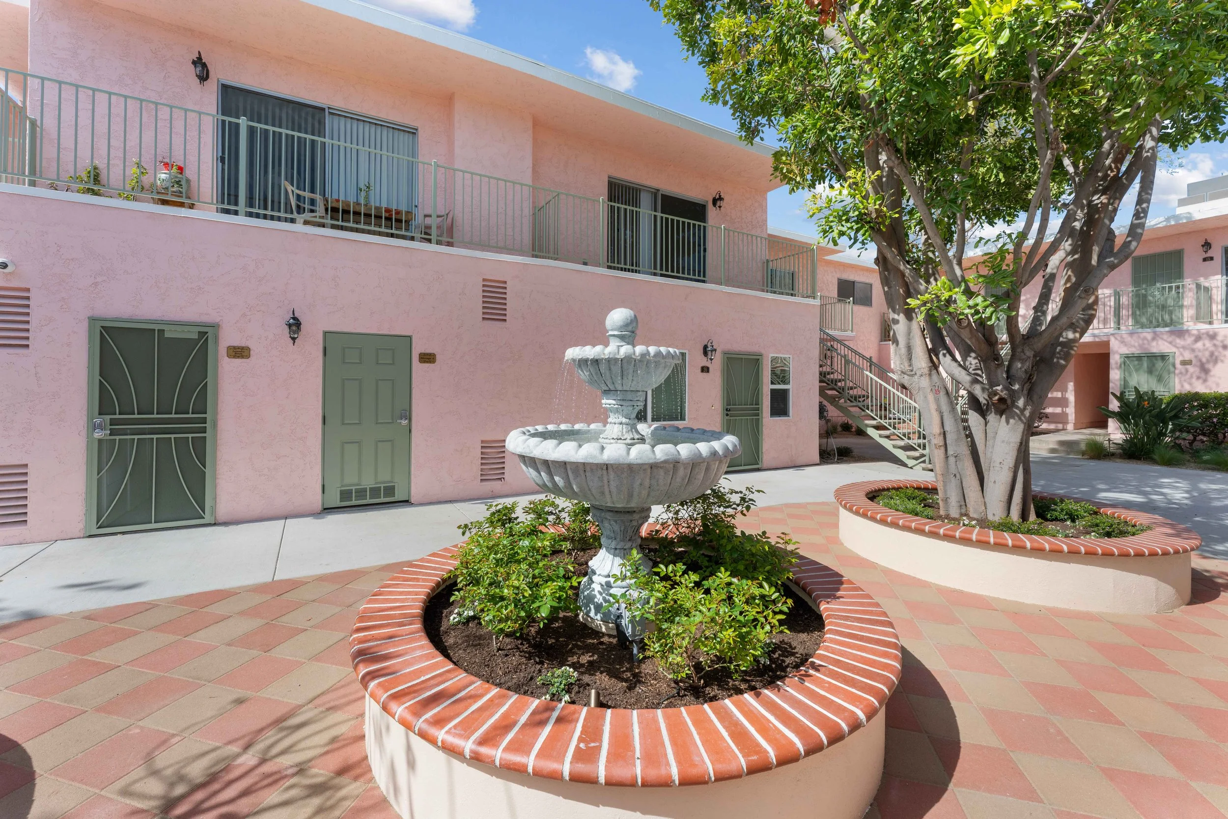 A pink building with glass doors and balconies, surrounded by a landscaped courtyard with a large tree, a decorative fountain, and paving stones.