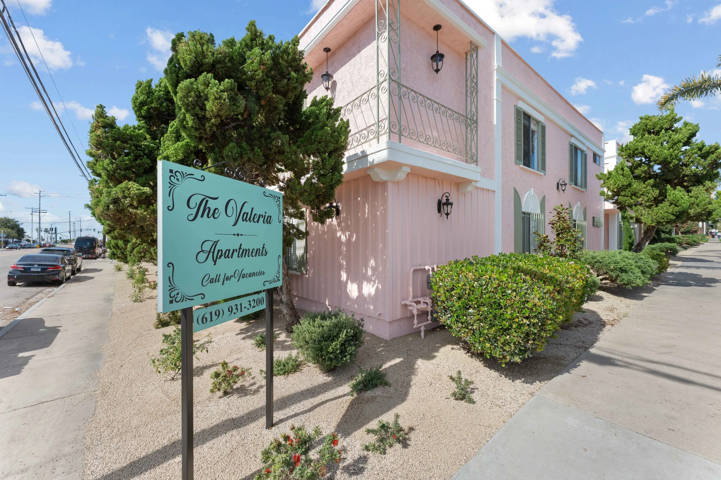 Pink two-story apartment building with a balcony, surrounded by trees and bushes; street with parked cars and a sign that reads "The Valeria Apartments Call for Vacancies" and a phone number.